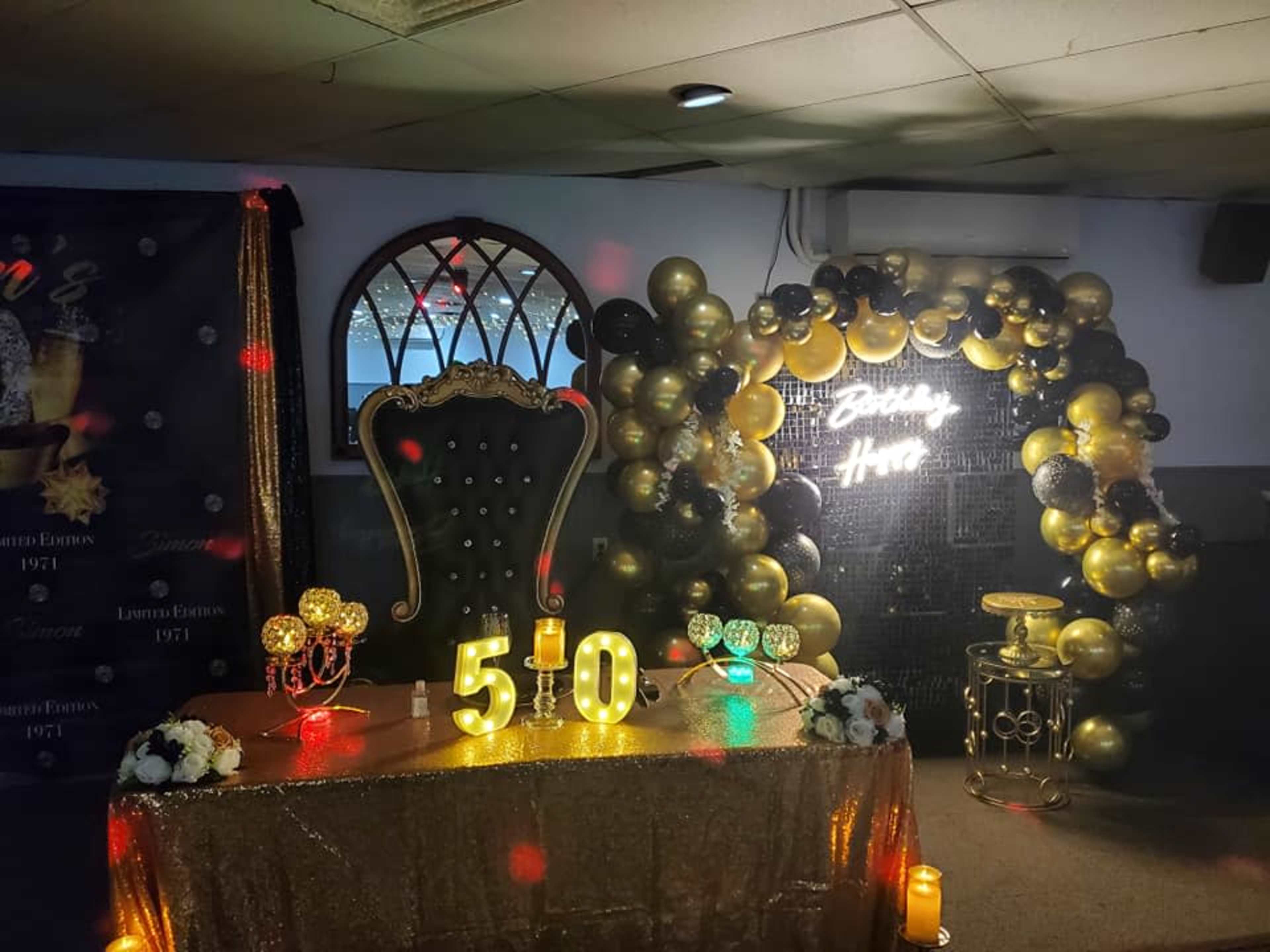 The image shows a decorated party setup featuring a throne-like chair, a table with the number "50" illuminated, and a balloon arch in black and gold surrounding a neon sign that says "Birthday Home."