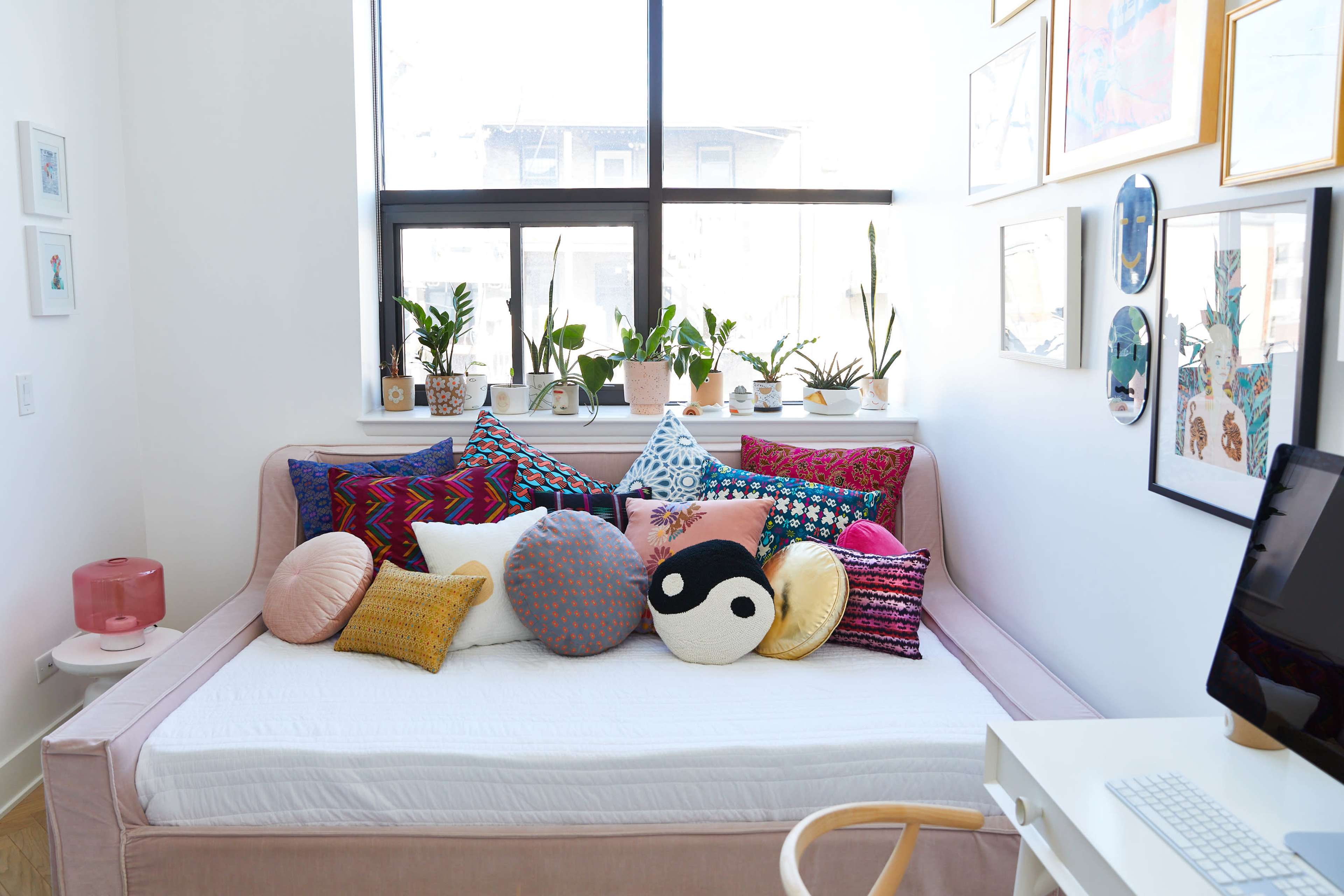 A bright room features a daybed adorned with various colorful pillows, surrounded by potted plants and framed artwork on the walls.