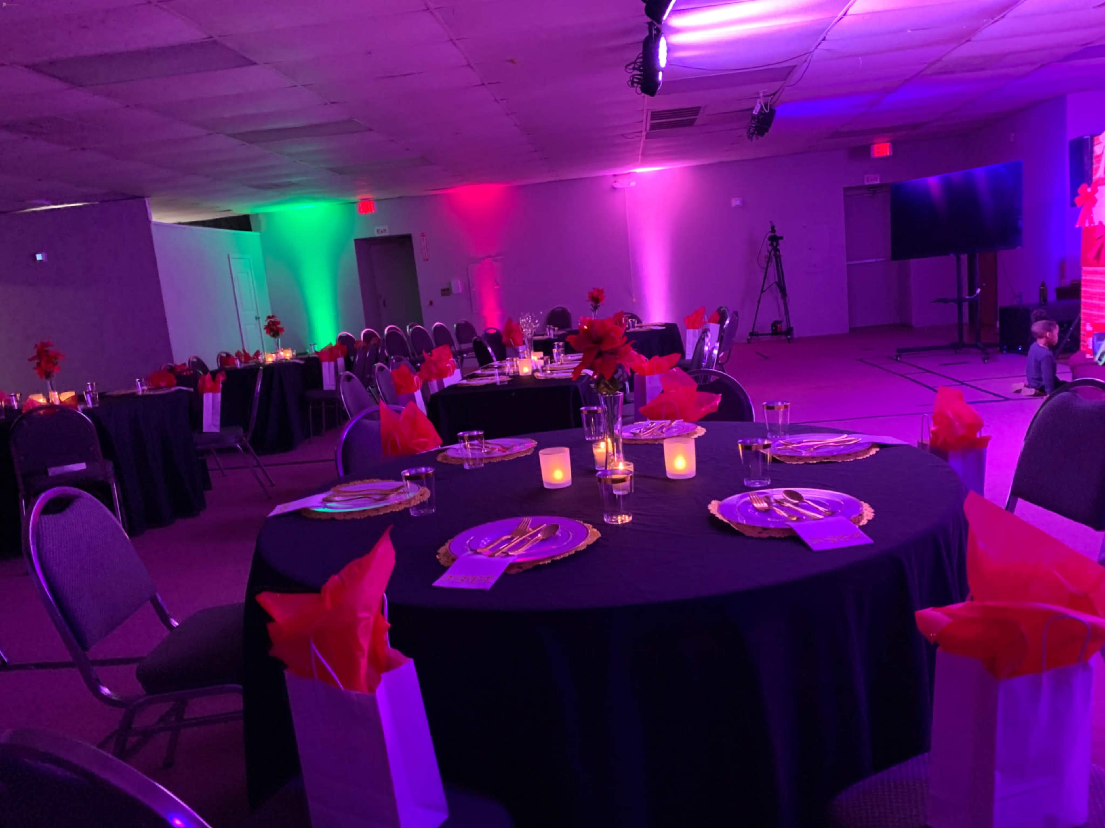 The image shows a banquet hall set up for an event, featuring round tables with black cloths, gold plates, and decorative centerpieces, illuminated by colorful lights.