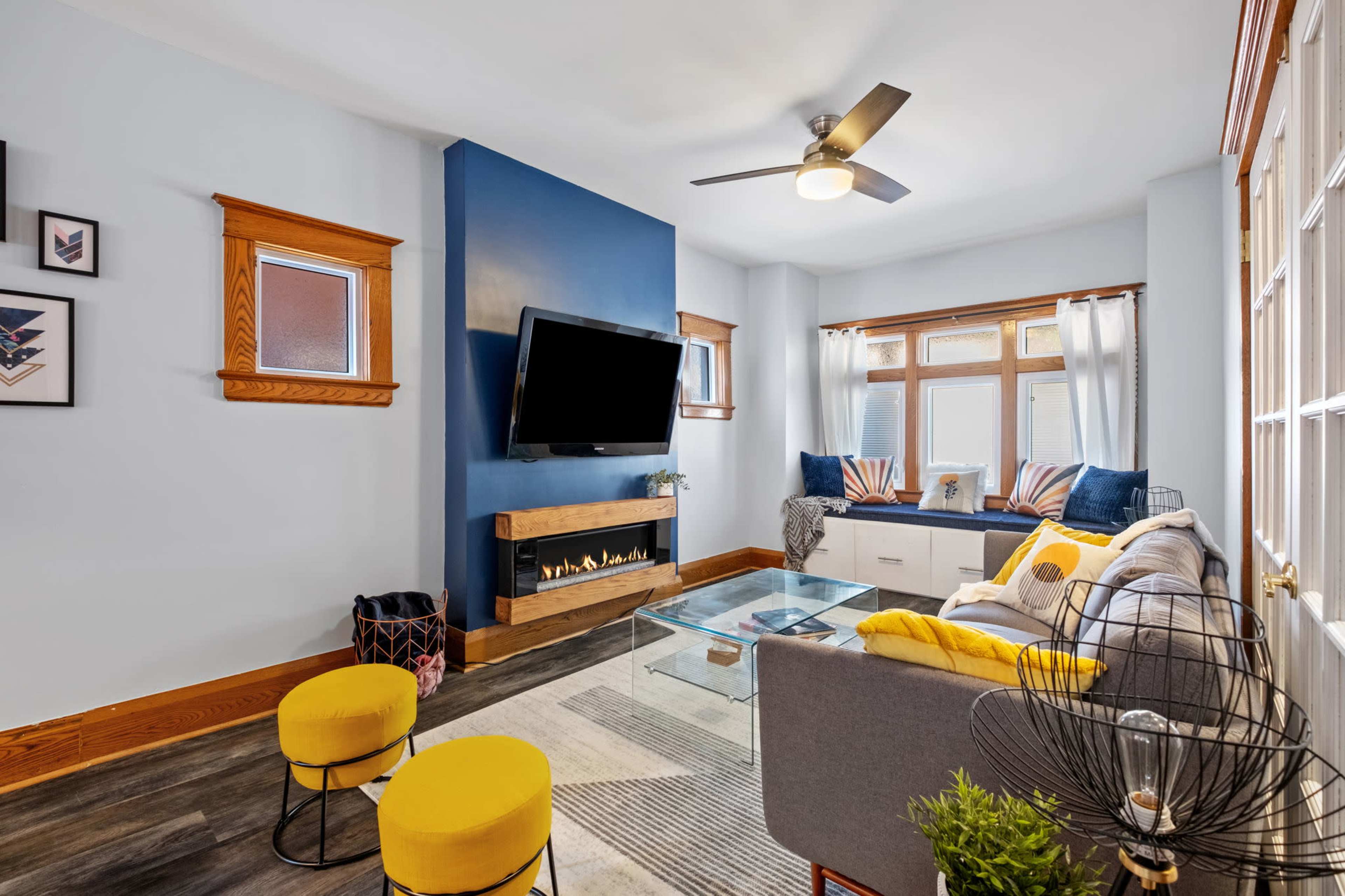 The image shows a modern living room featuring a mounted television, a glass coffee table, a cozy seating area with yellow accent stools, and a window nook with cushions.