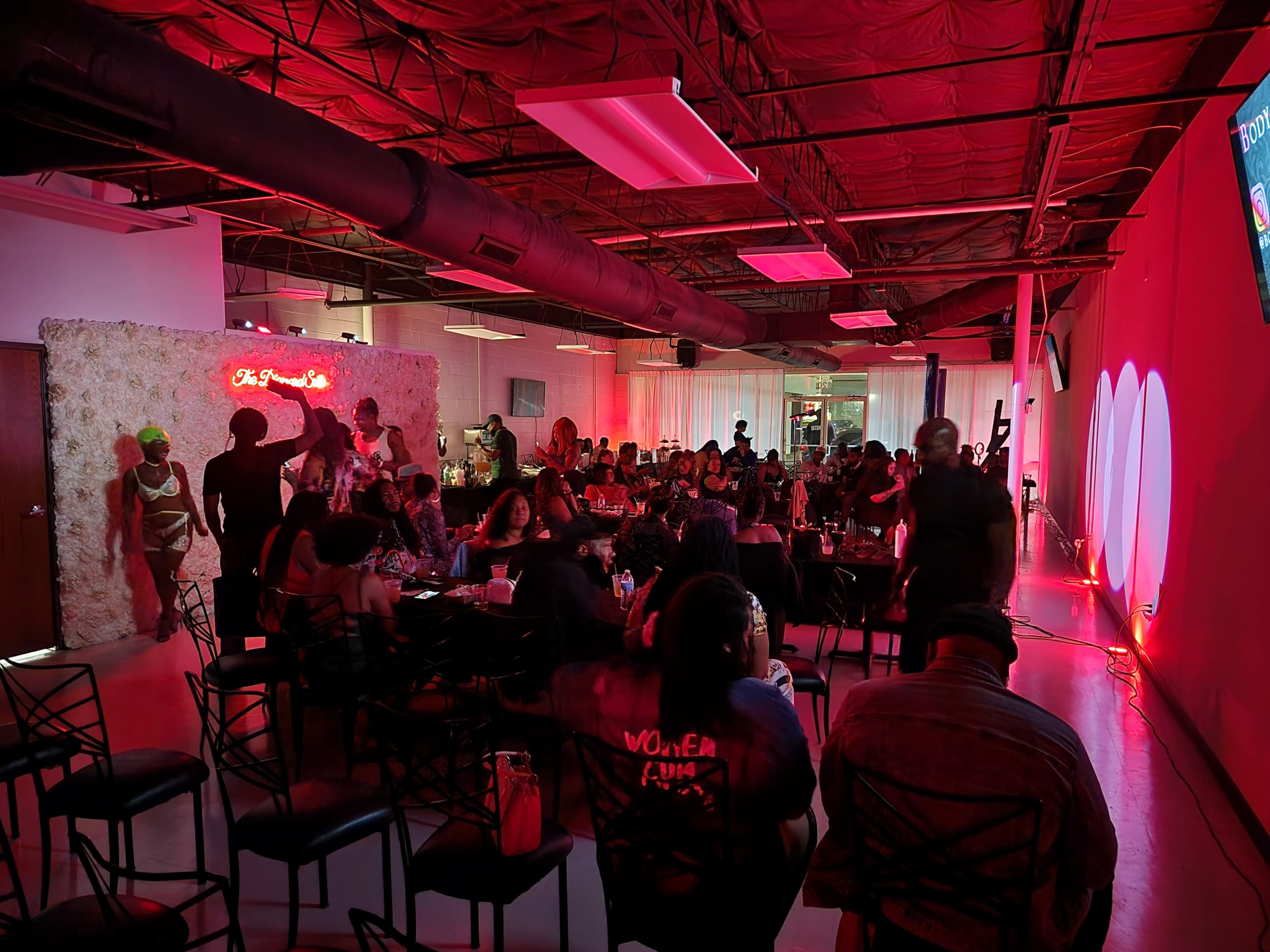 A lively indoor event space features a gathering of people seated and mingling, with red lighting and a decorative wall in the background.
