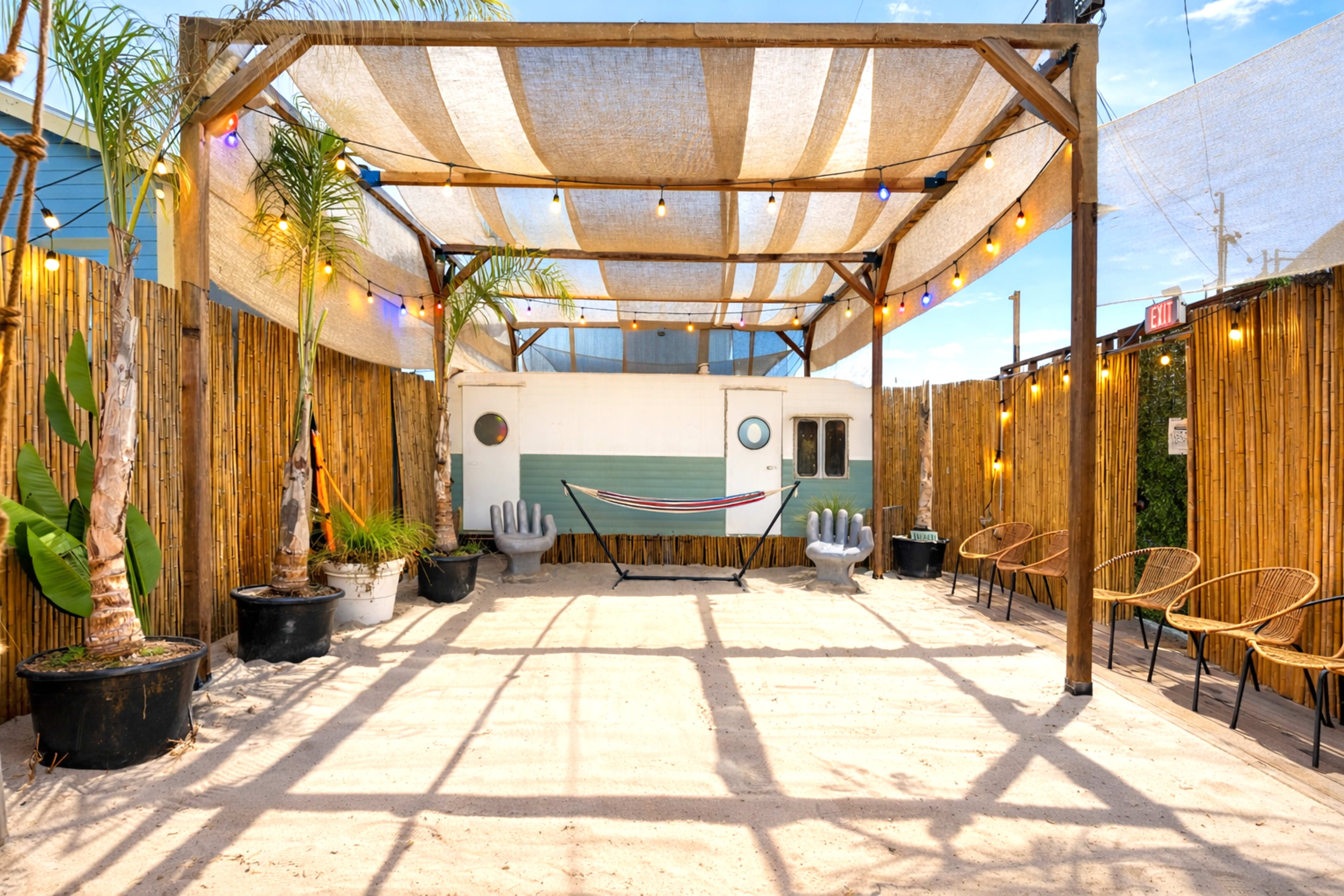 The image shows an outdoor space with a wooden structure covered by a striped canopy, featuring potted plants, sandy ground, and cozy seating arrangements.