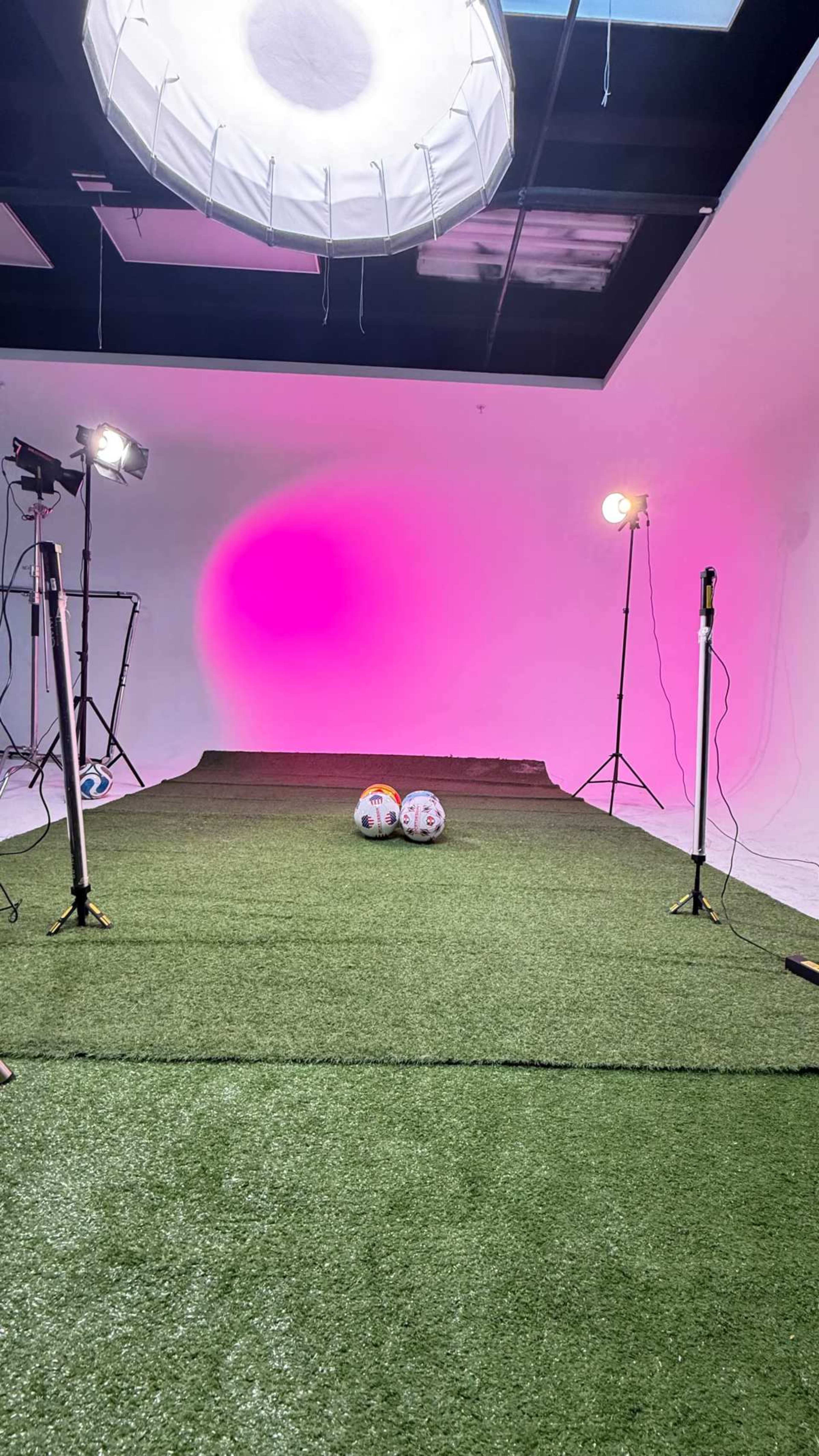 The image shows a studio setup for a photoshoot with artificial grass, two soccer balls, and multiple lighting equipment creating a colorful backdrop.