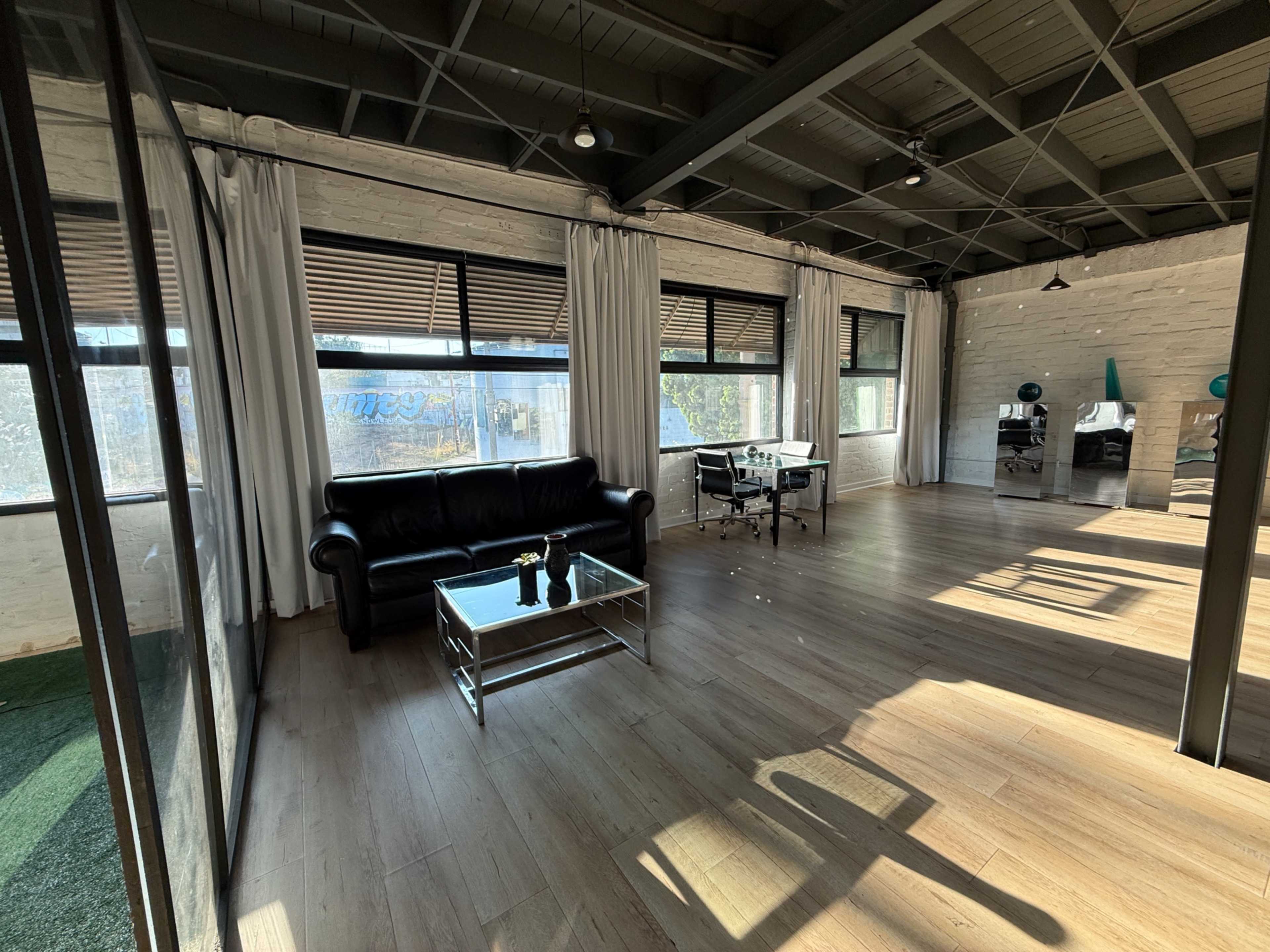 The image shows a spacious office interior with a black leather couch, a glass coffee table, and a desk with a chair, all illuminated by natural light streaming through large windows.