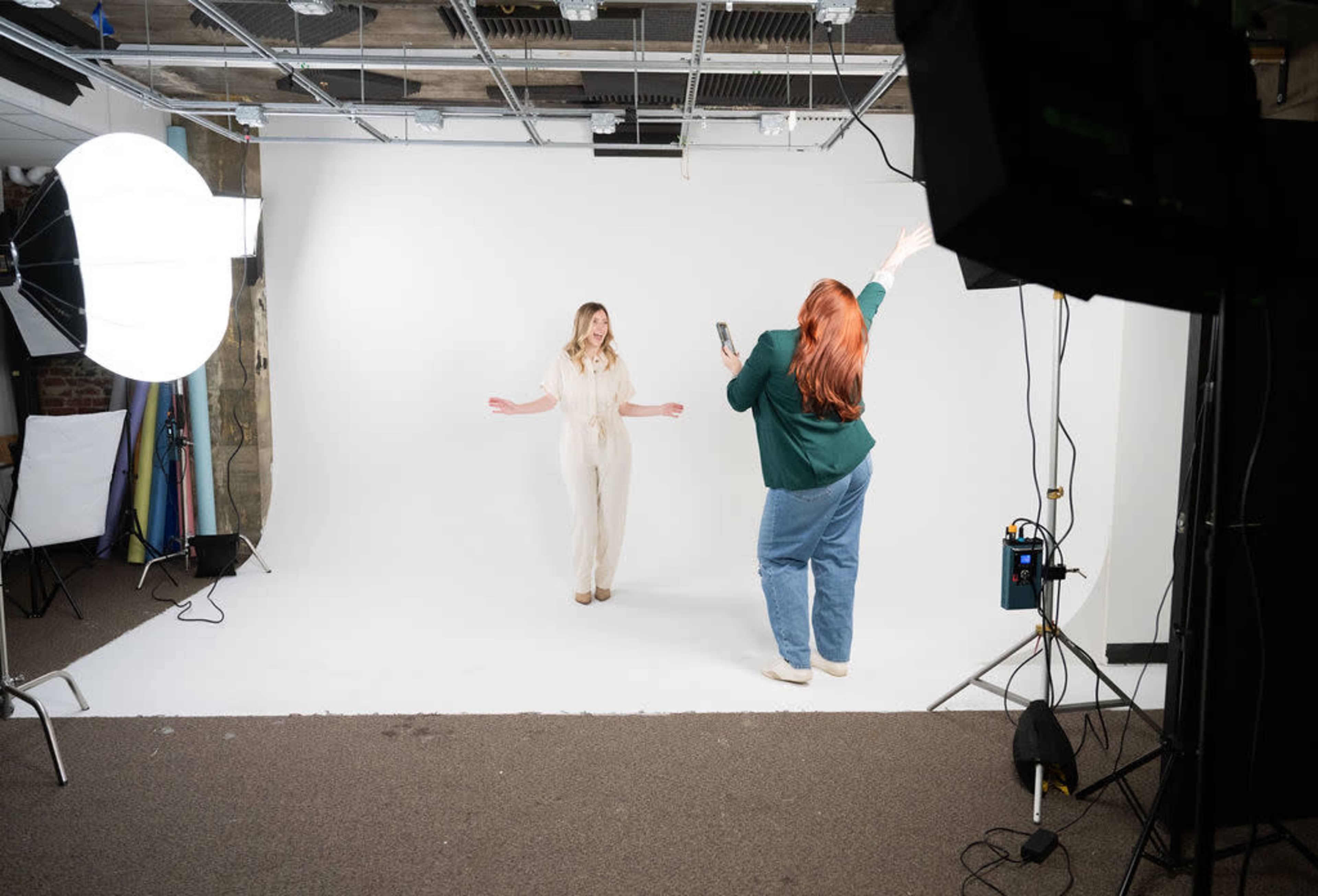 Two women are in a photography studio, one posing against a white backdrop while the other captures the moment with a camera.