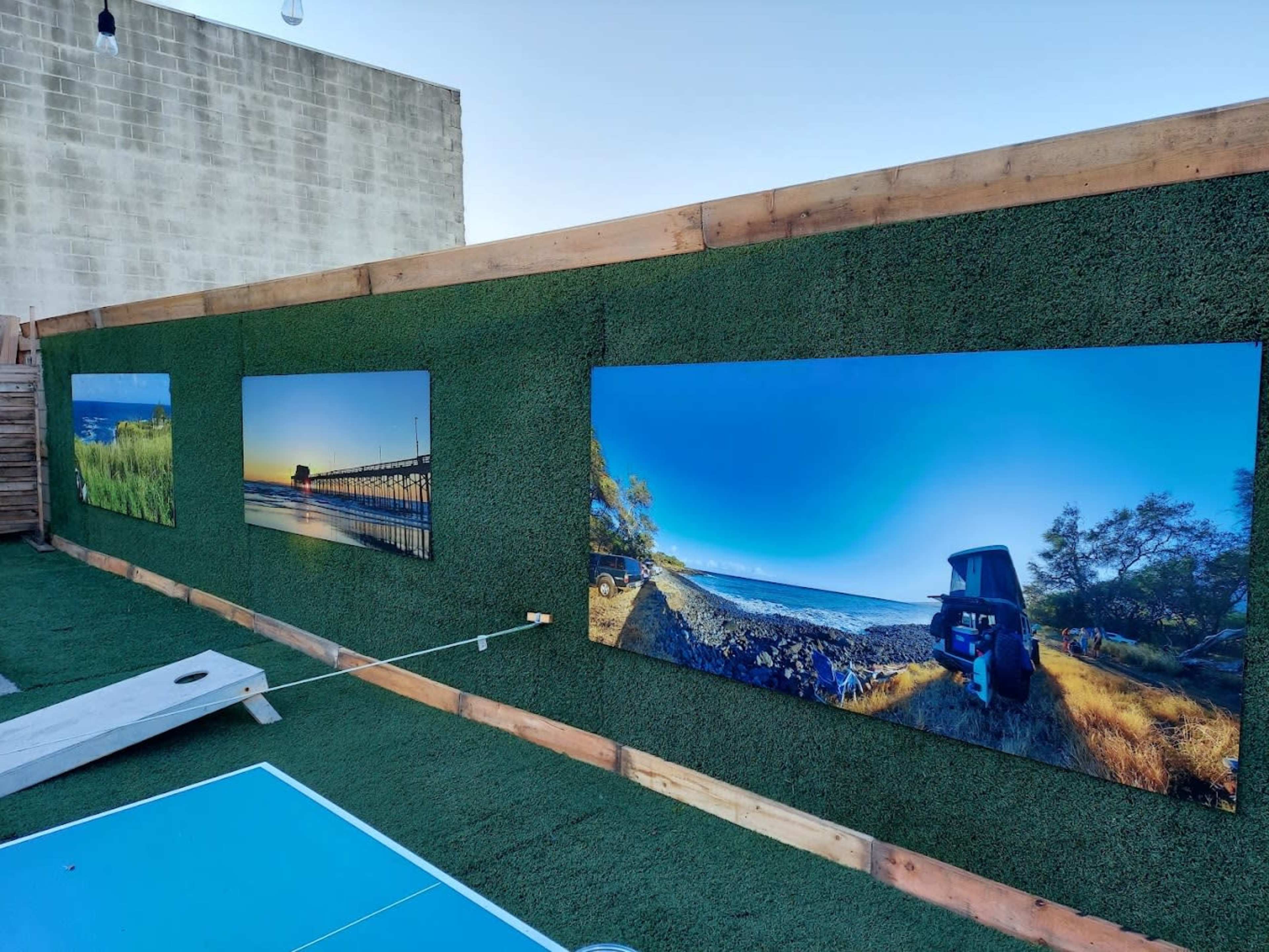 The image shows a green wall adorned with three large landscape photographs, featuring scenes of a pier, a coastline, and a vehicle near a beach, in an outdoor setting.