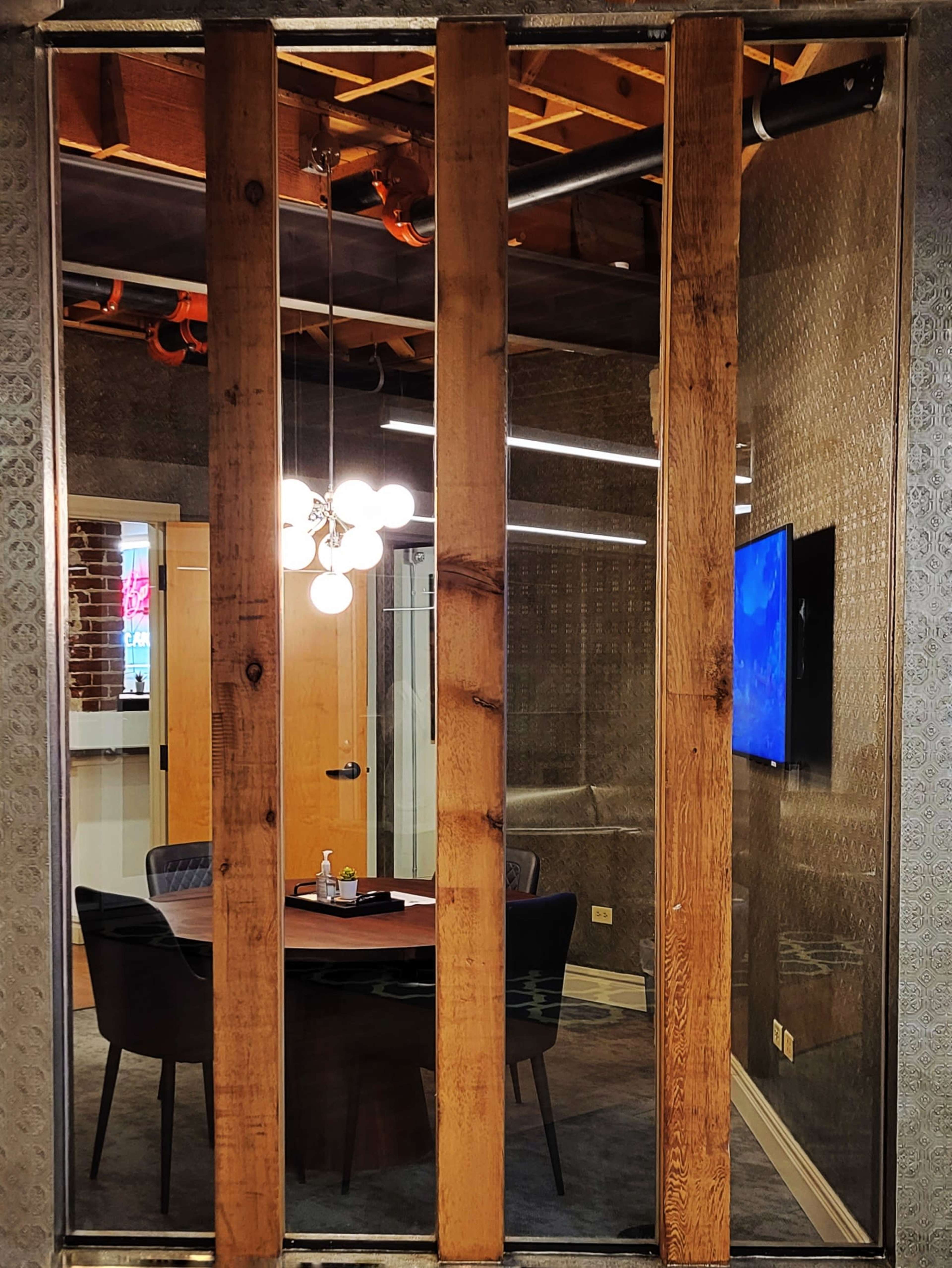 A modern conference room with a round table, several chairs, and a wall-mounted television, enclosed by glass panels and framed by wooden beams.
