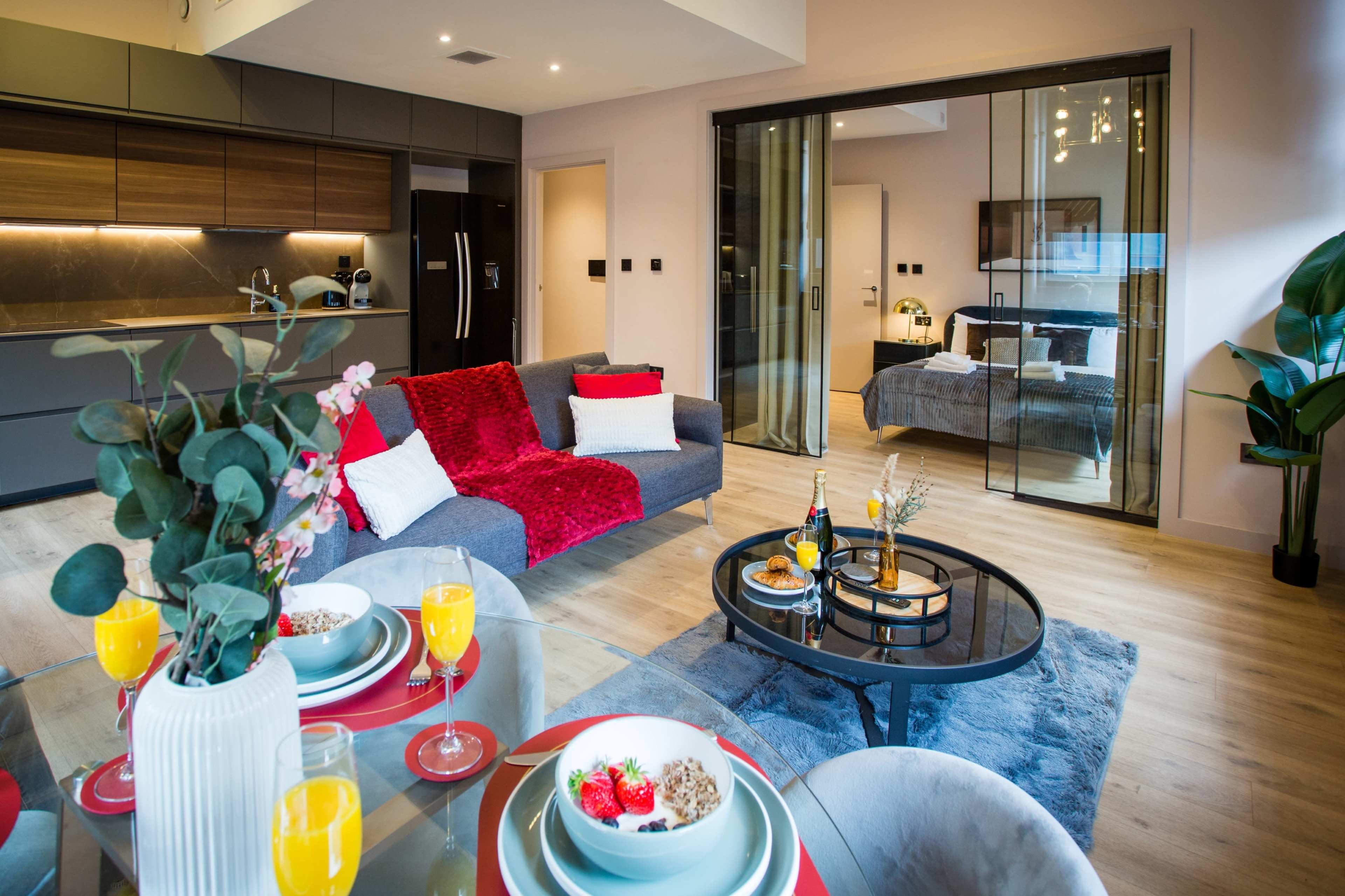 A modern living area features a gray sofa with a red throw, a glass dining table set for breakfast, and a visible bedroom through sliding glass doors.