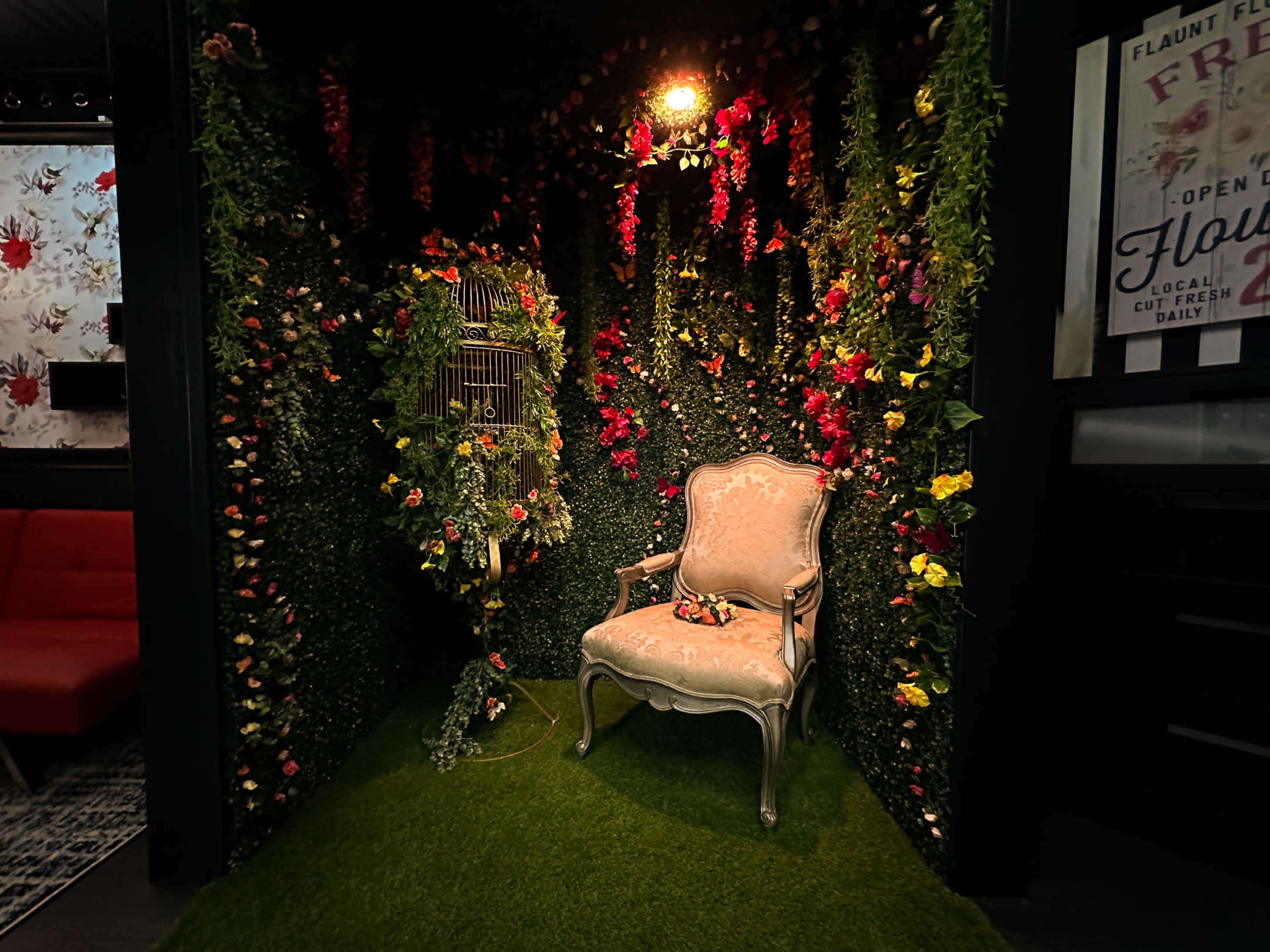 The scene features a vintage chair next to a decorative birdcage, surrounded by a wall of vibrant artificial plants and flowers.