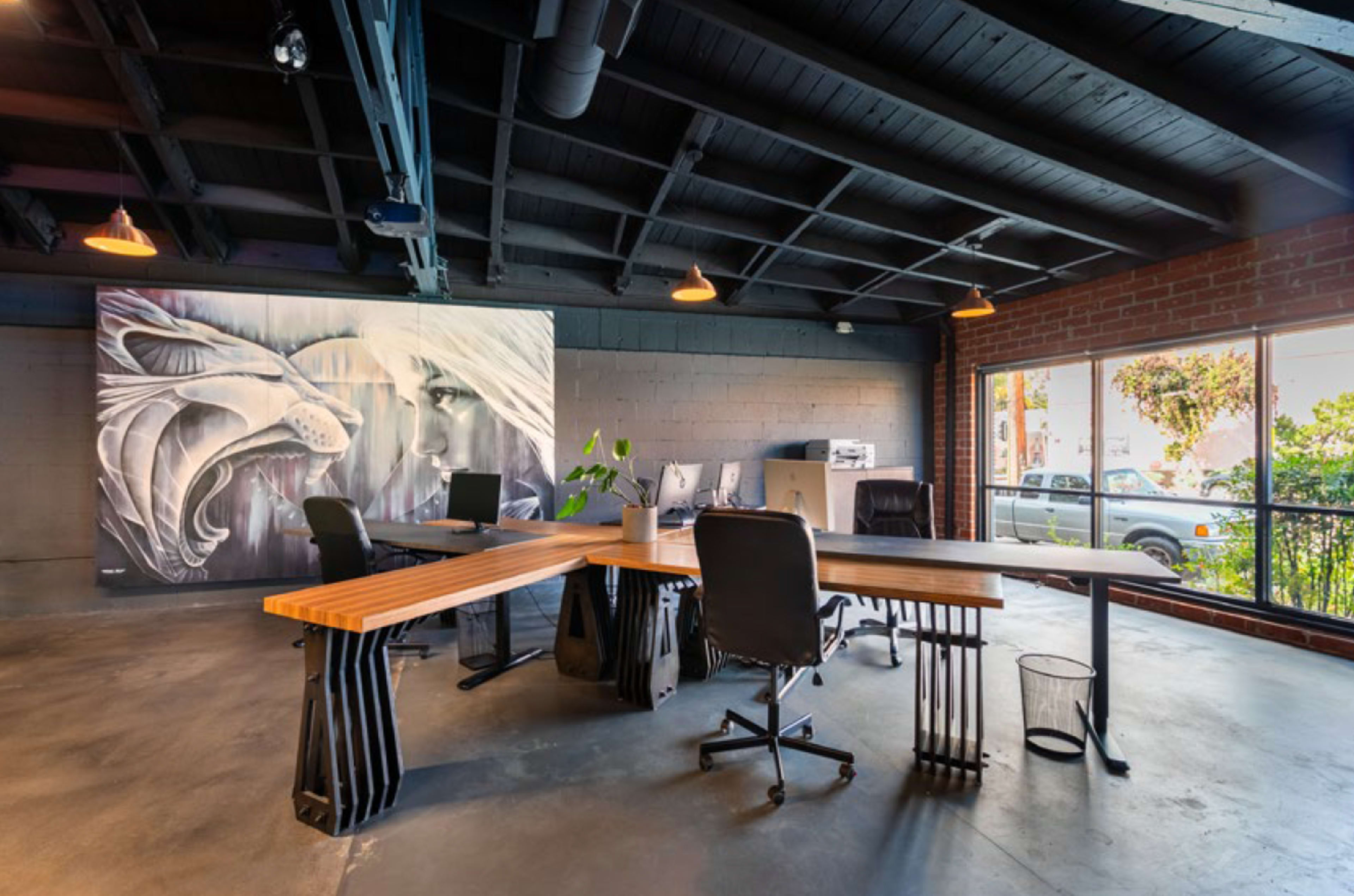 A modern office space features a large wooden table surrounded by black chairs, with a mural of a lion and a woman's face on the wall.