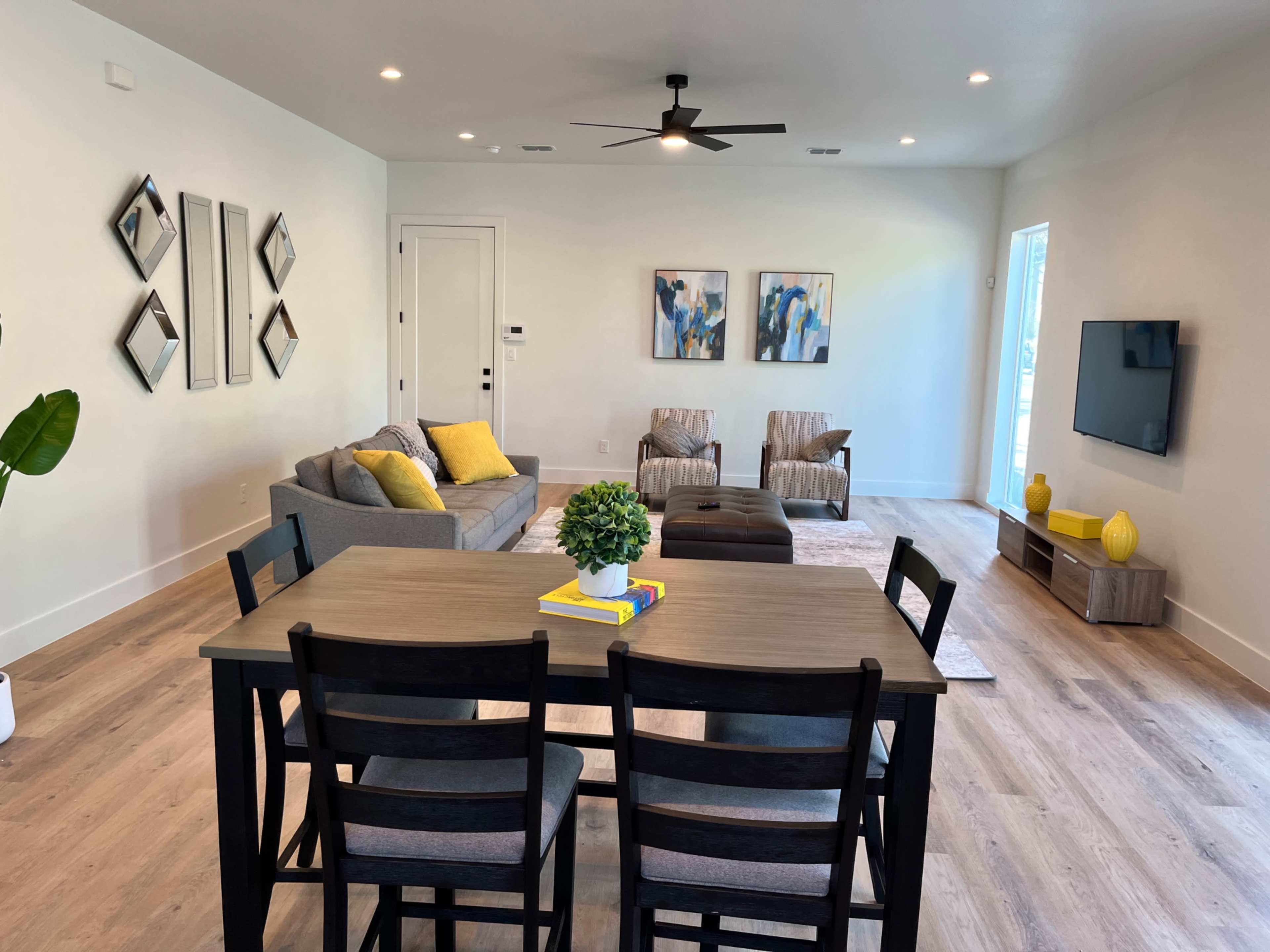 The room features a dining area with a table and chairs, a living space with a sofa and armchairs, and a media console, all illuminated by overhead lighting.