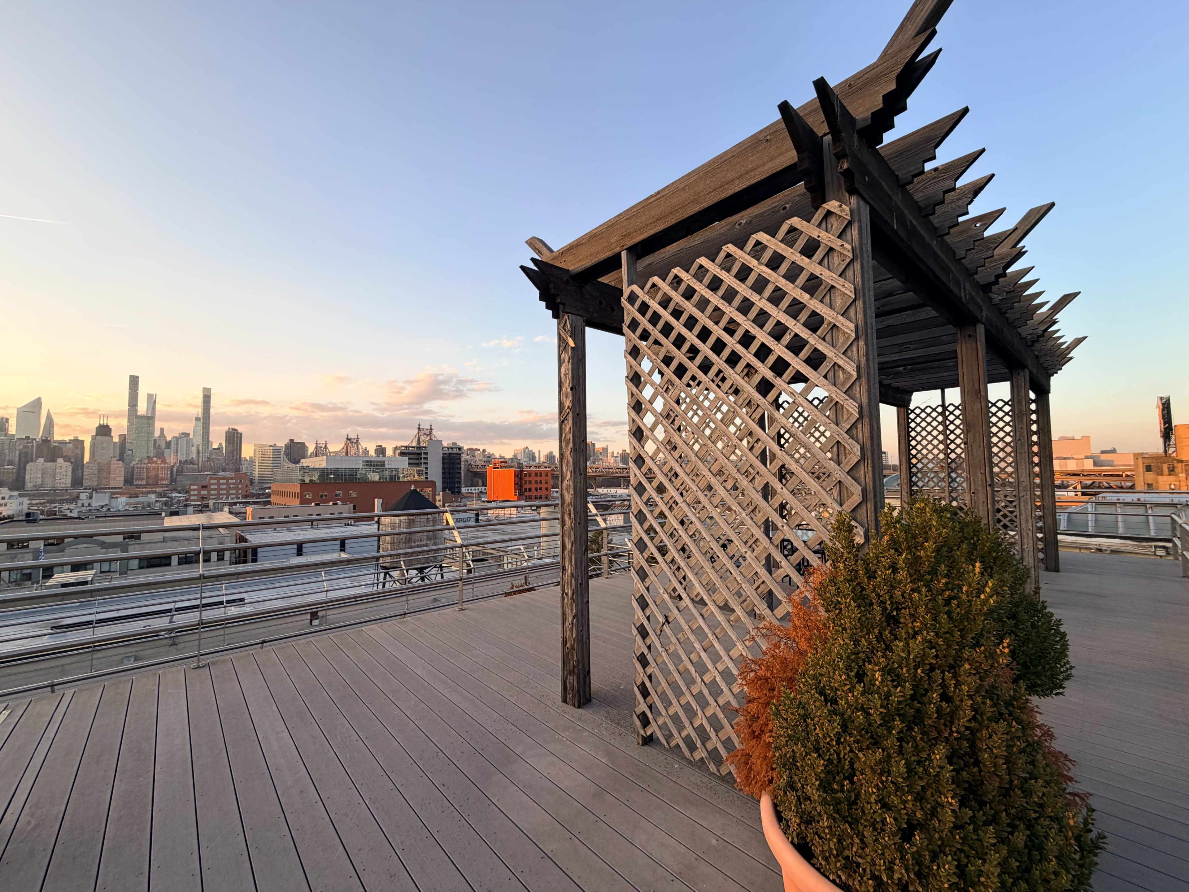 A wooden pergola with a lattice design stands on a rooftop overlooking a city skyline at sunset.