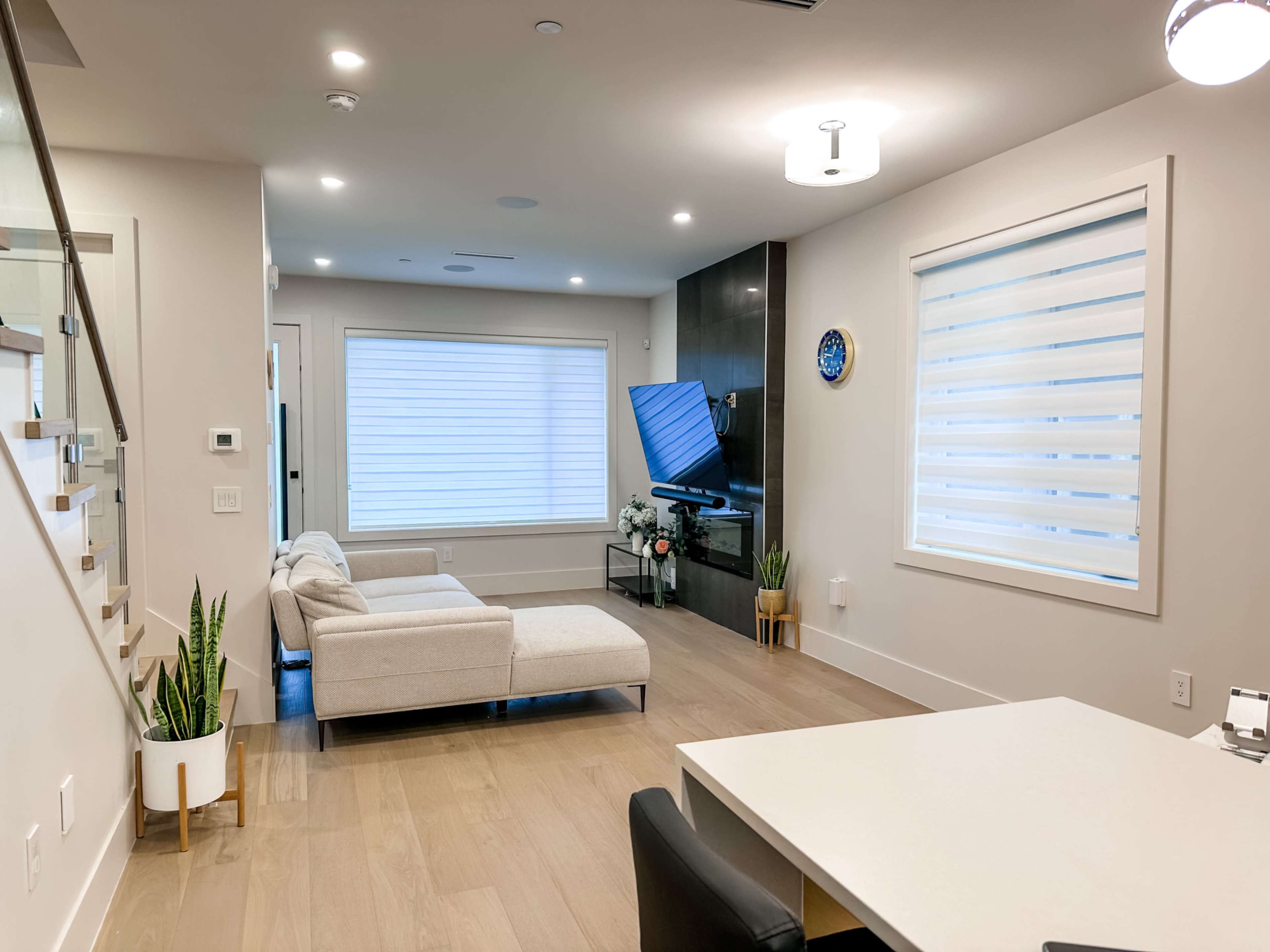 A modern living room featuring a light-colored sectional sofa, a television on a dark wall, and a dining area with a white table and black chairs.