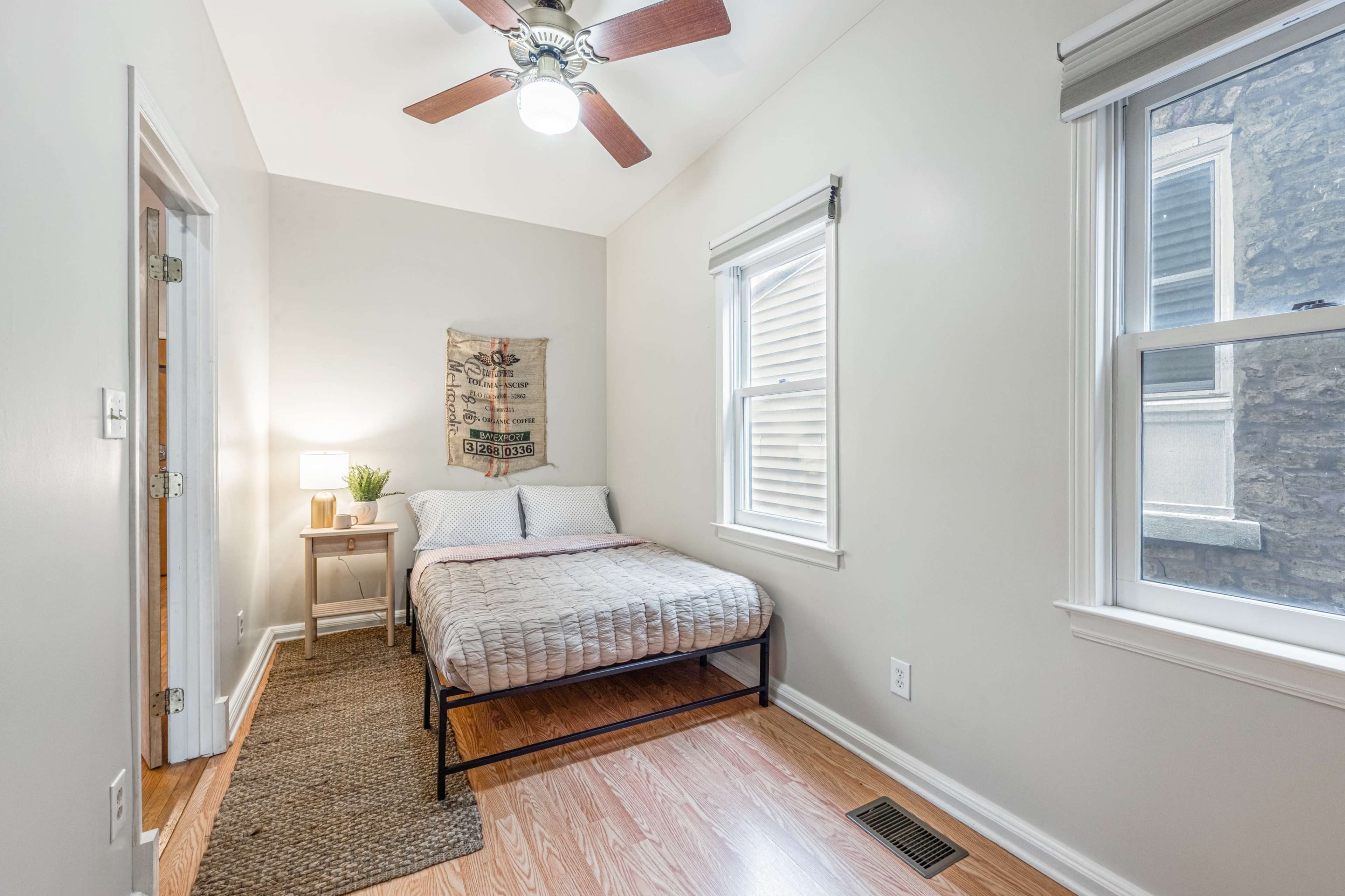 The image shows a small bedroom featuring a bed, a nightstand with a lamp, and two windows, all set against light-colored walls and a wooden floor.