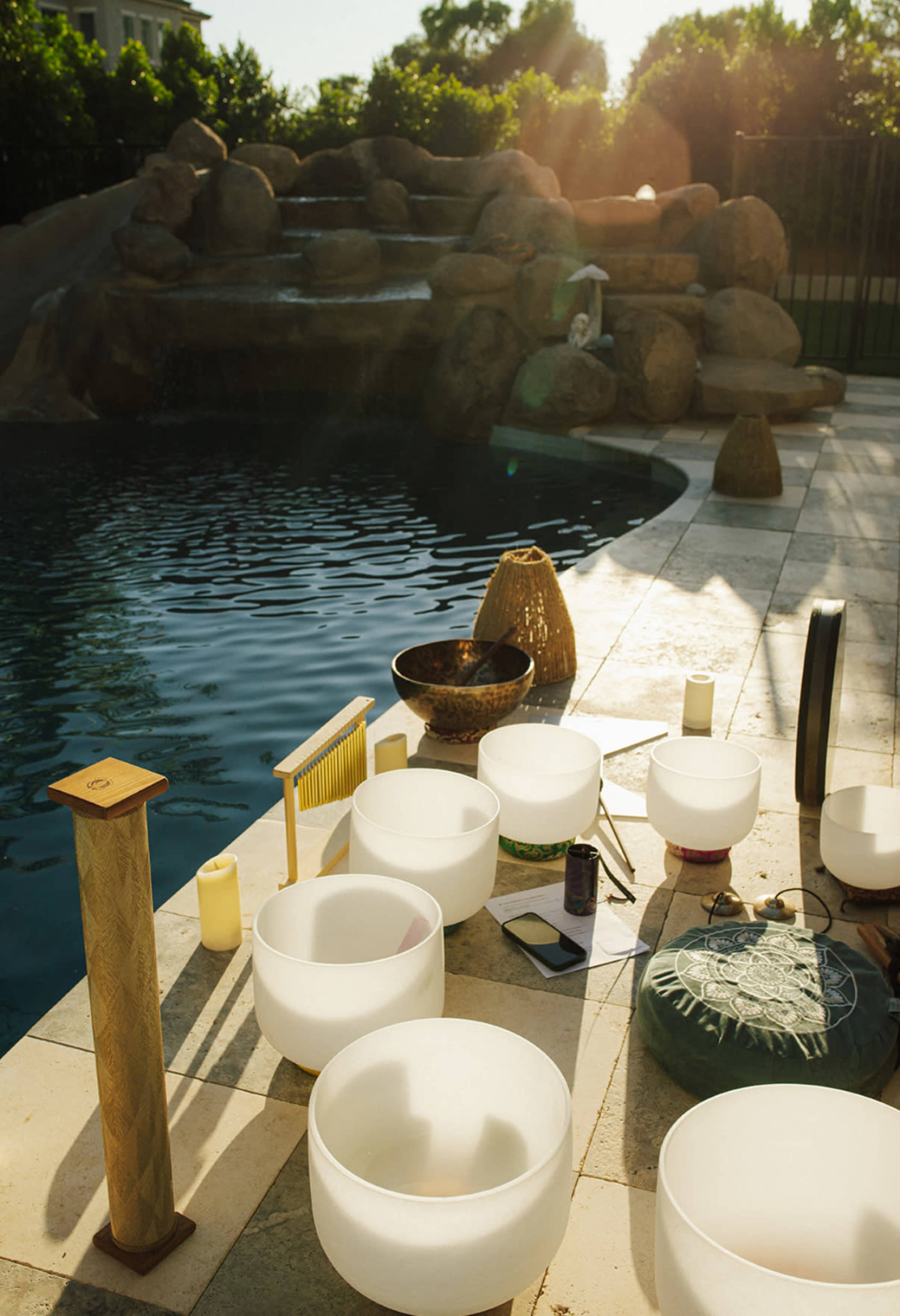 The scene features a collection of crystal singing bowls arranged on a stone patio beside a swimming pool, with a rocky waterfall in the background and sunlight reflecting off the water.