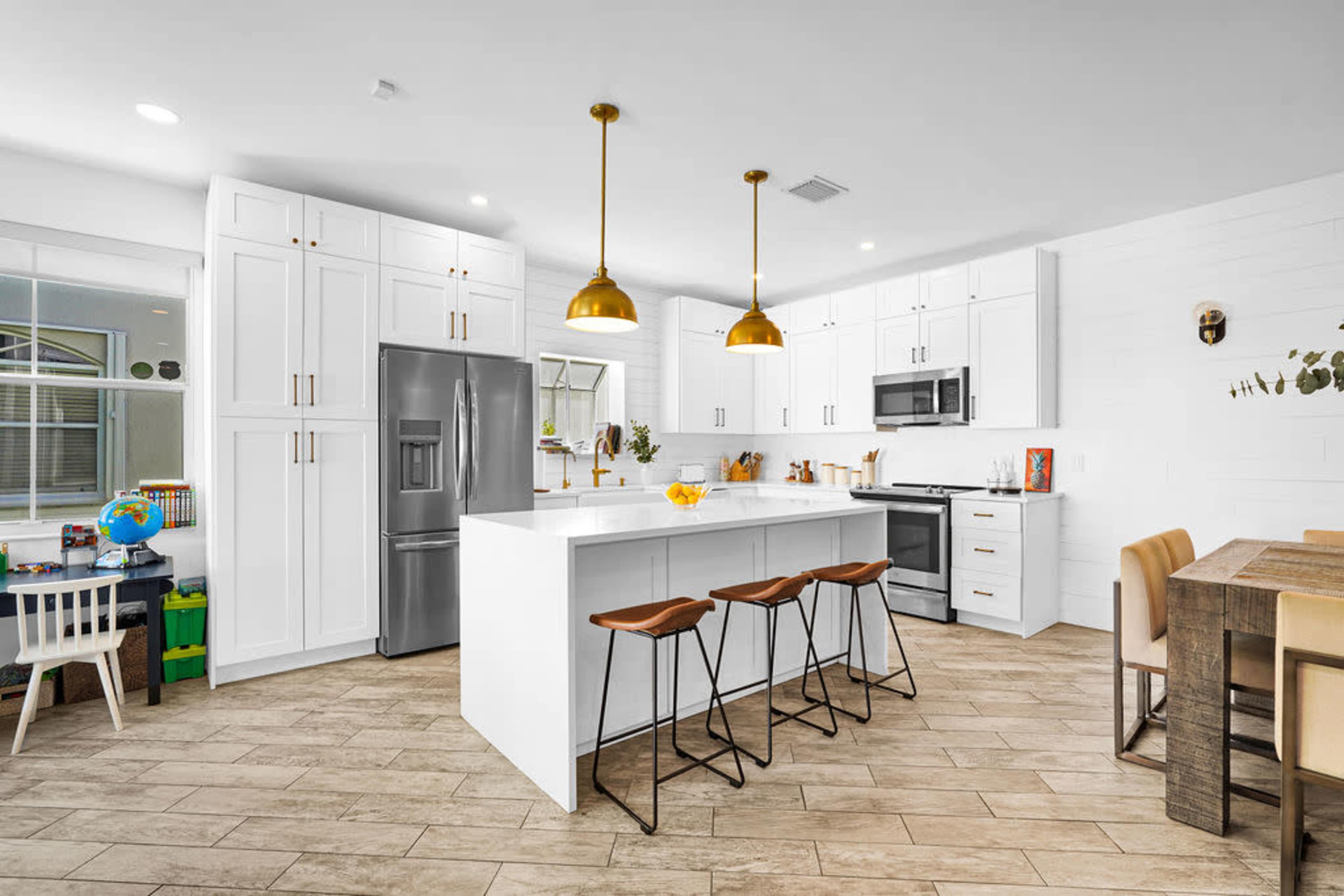 A modern kitchen features white cabinetry, stainless steel appliances, a central island with seating, and pendant lighting.