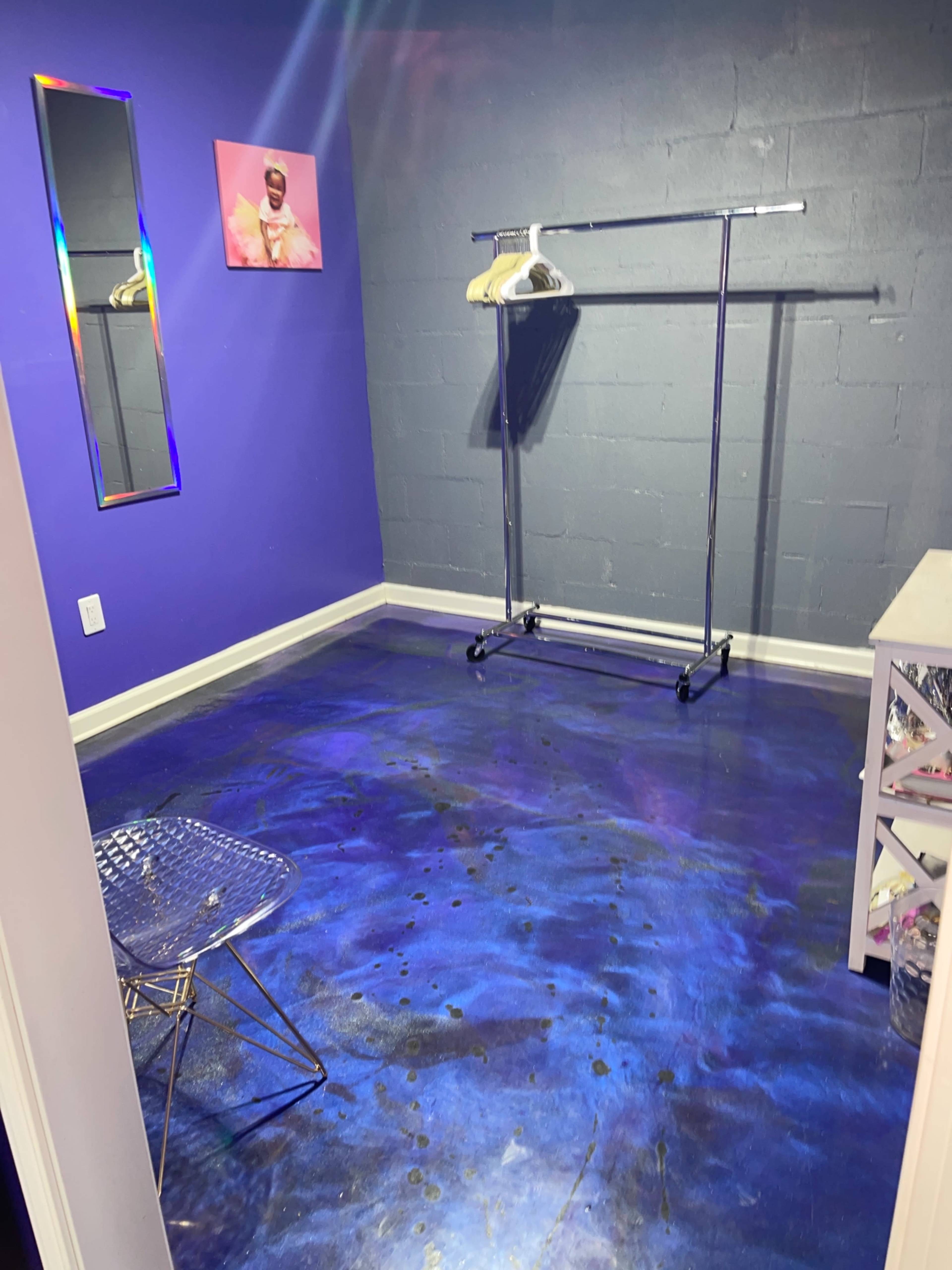 A small room with purple walls, a clothing rack, a mirror, and a metallic chair on a colorful floor.