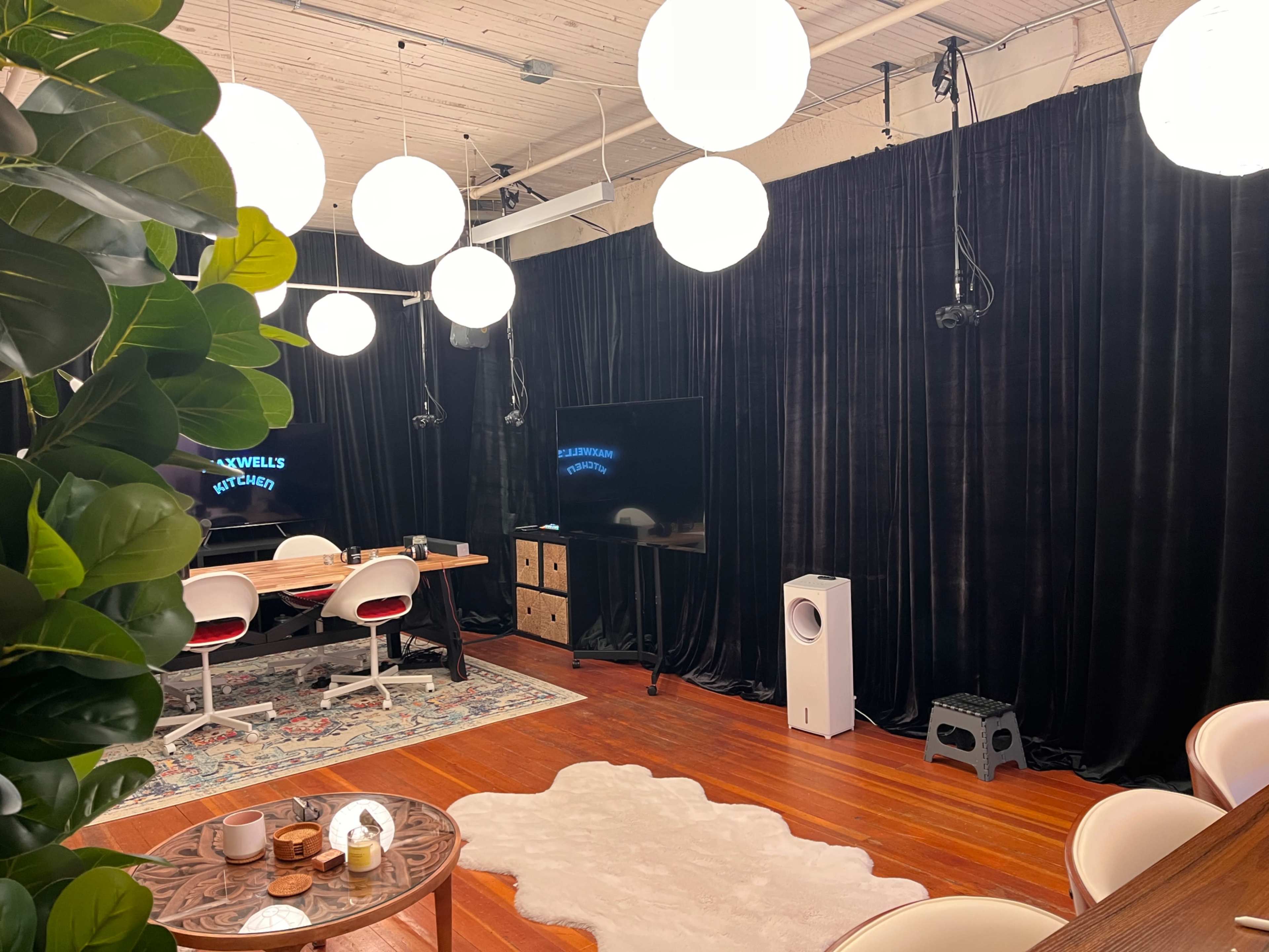 The image shows a staged room with black curtains, a large screen, a dining table with chairs, and decorative lighting fixtures.