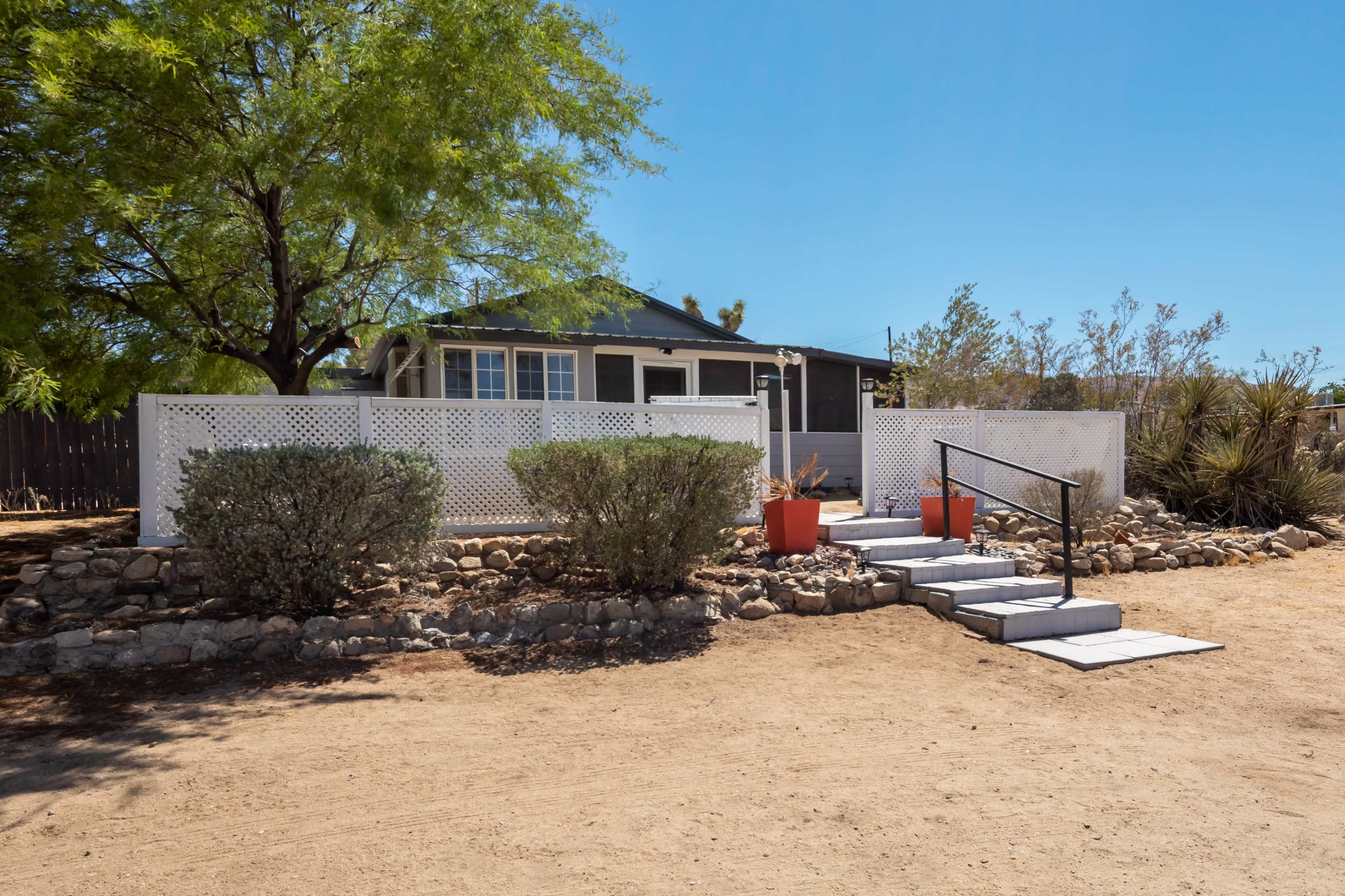 A single-story house with a porch is surrounded by low white fencing, a gravel path, and potted red plants.