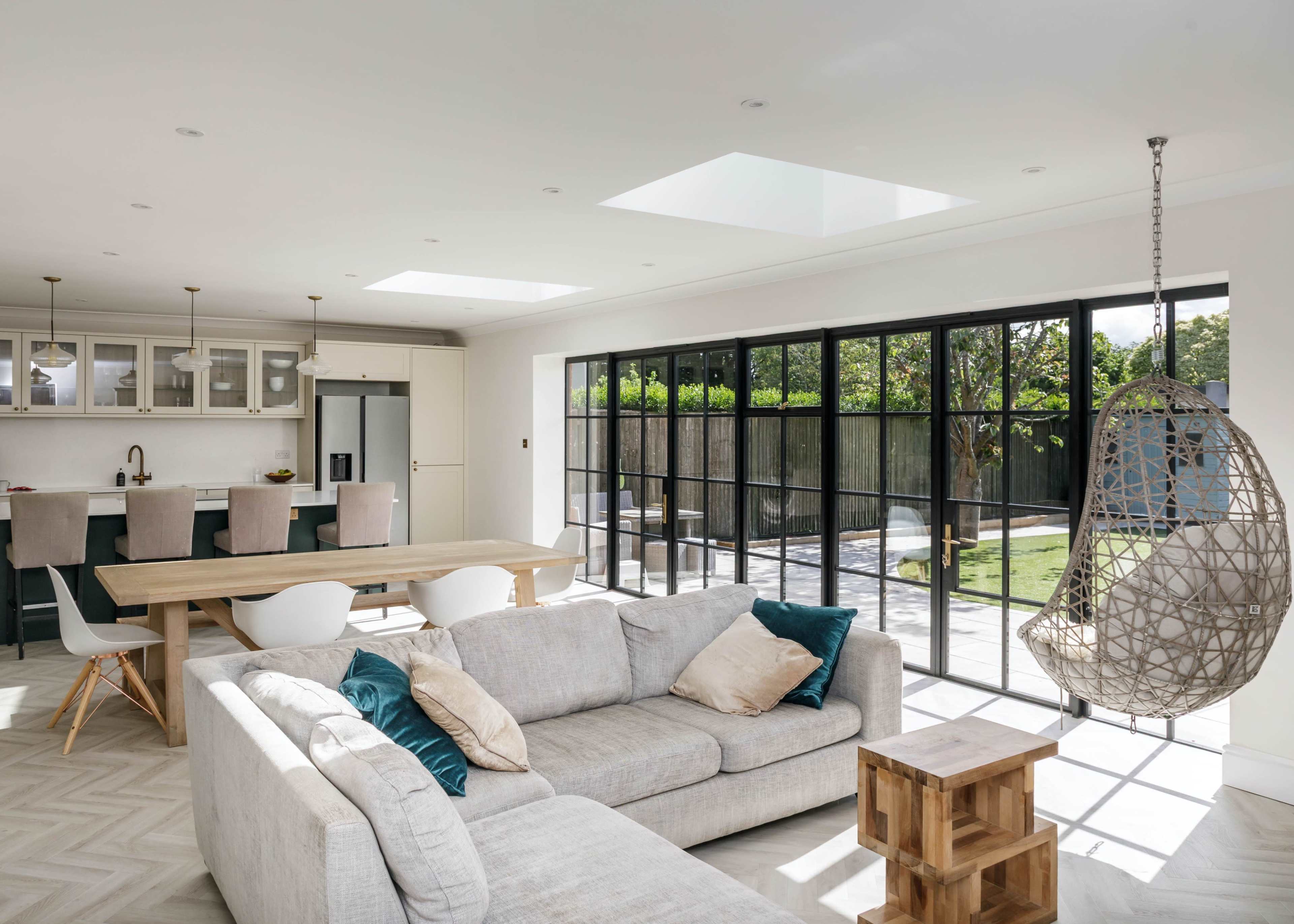 The image shows a modern living room with a light gray sectional sofa, a wooden dining table, a hanging chair, and large glass doors that open to a backyard.