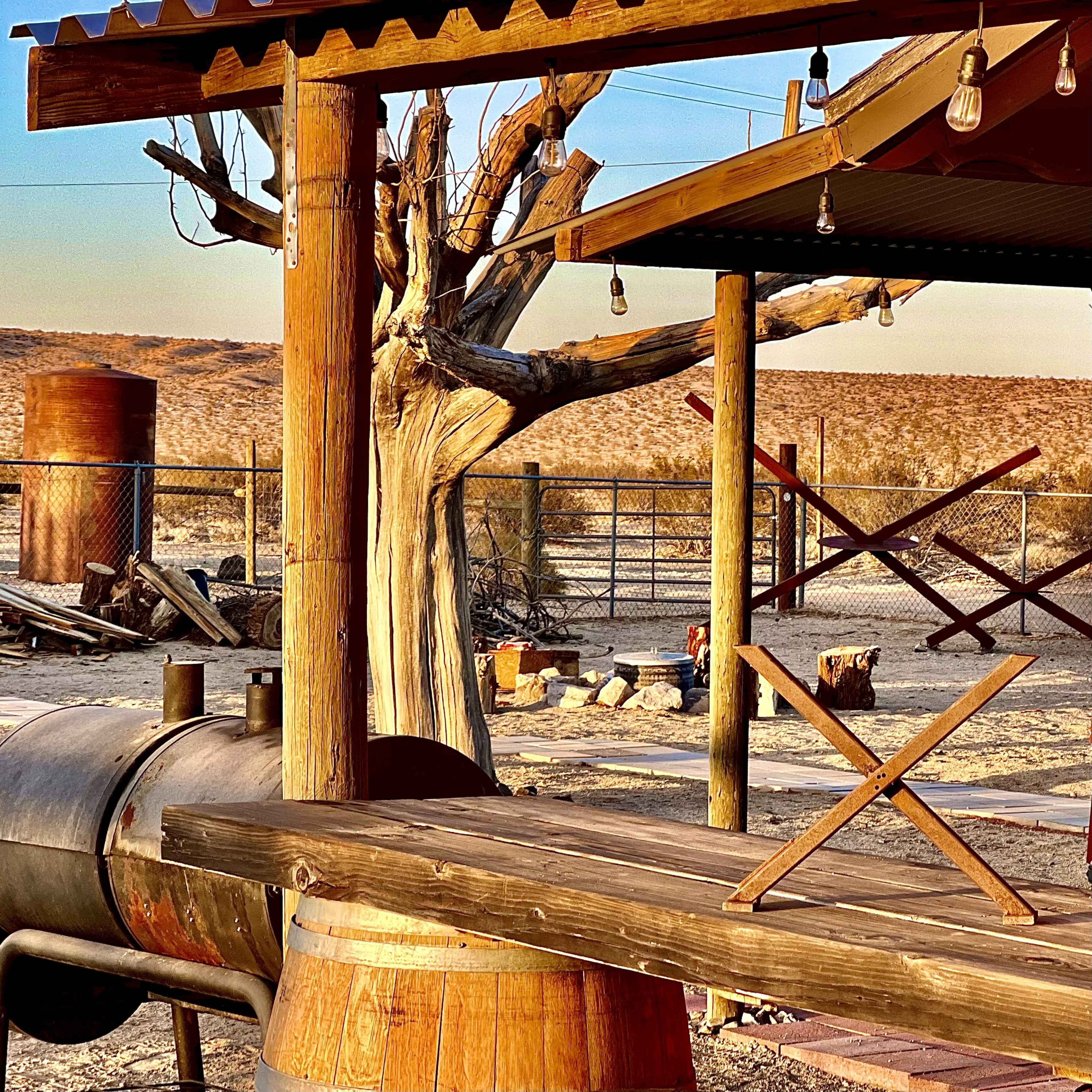 The image shows a rustic wooden structure near a metal barbeque grill and a barren tree, with a fenced landscape in the background.