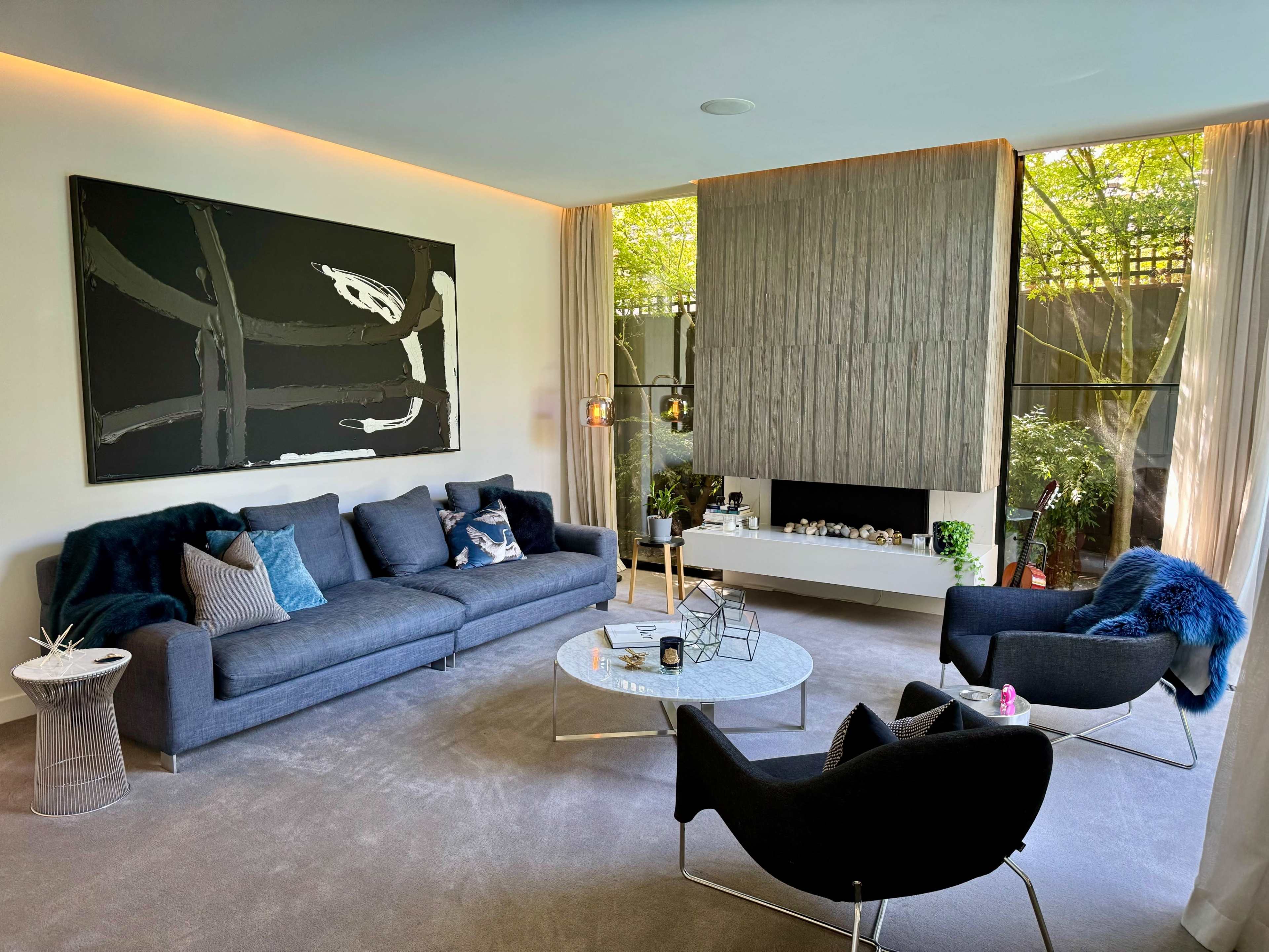 A modern living room features a gray sofa, a glass coffee table, and stylish armchairs, surrounded by large windows that allow natural light and greenery to enter.