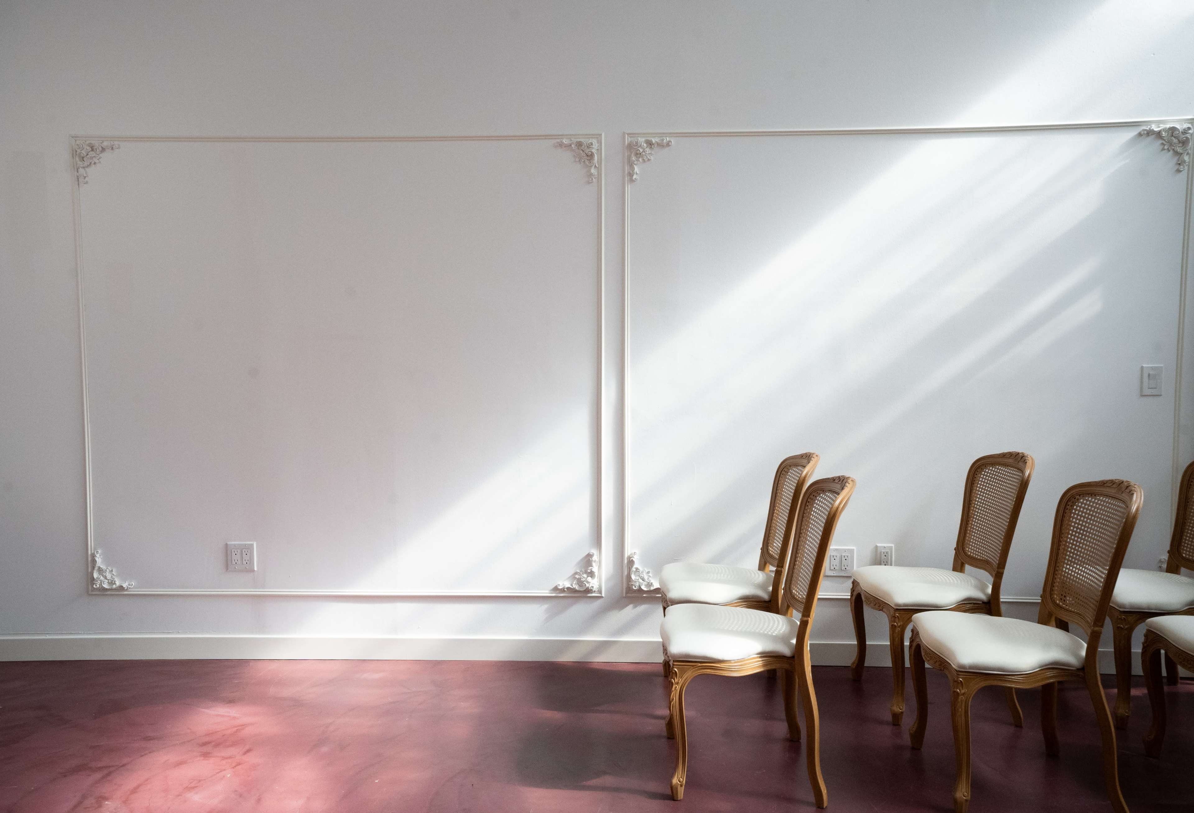The image shows a bright room with two empty wall frames and a row of wooden chairs with light-colored cushions arranged against one wall.
