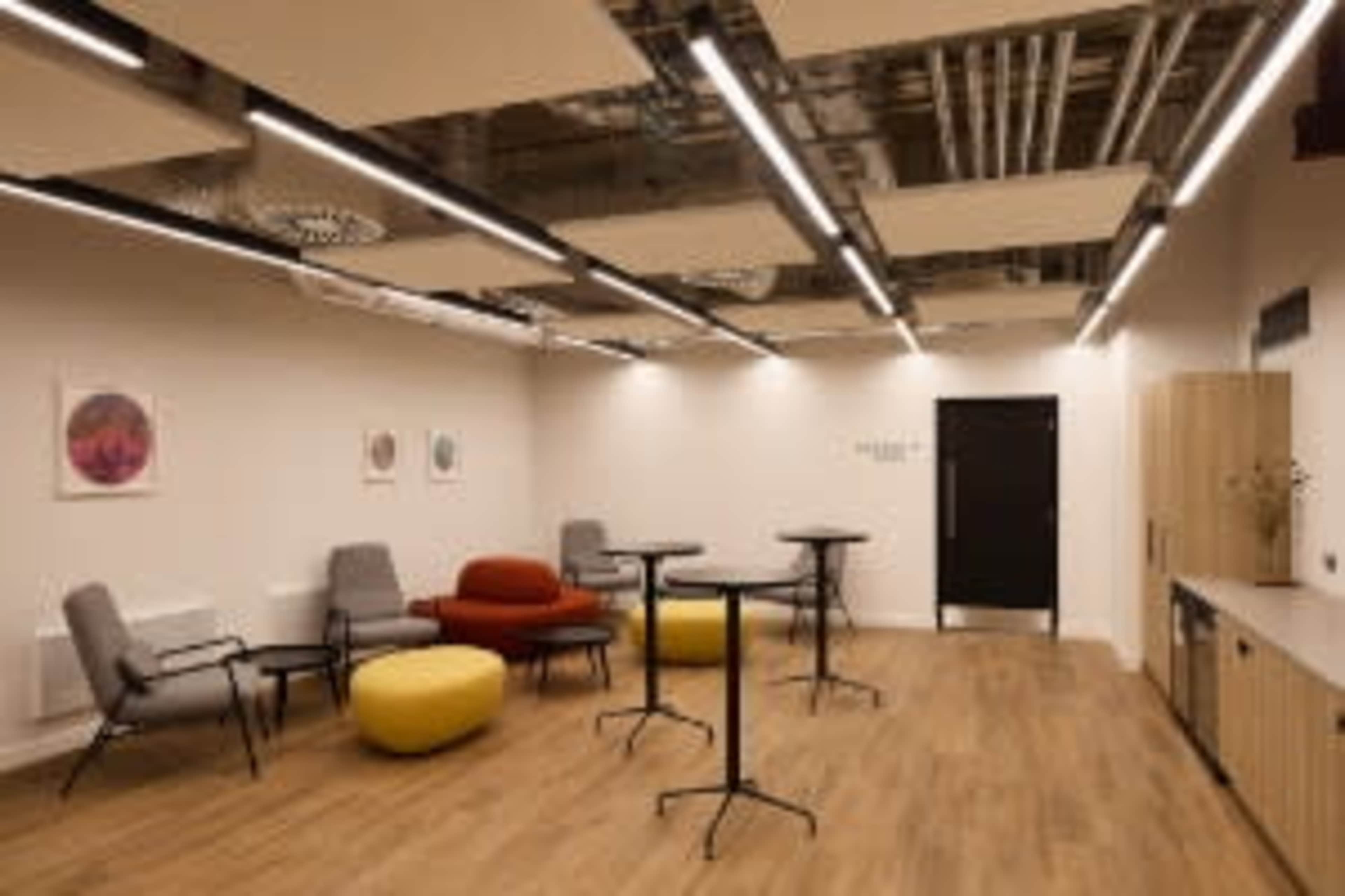 The image shows a modern meeting room featuring several chairs, small tables, and a kitchenette area with wooden cabinets.