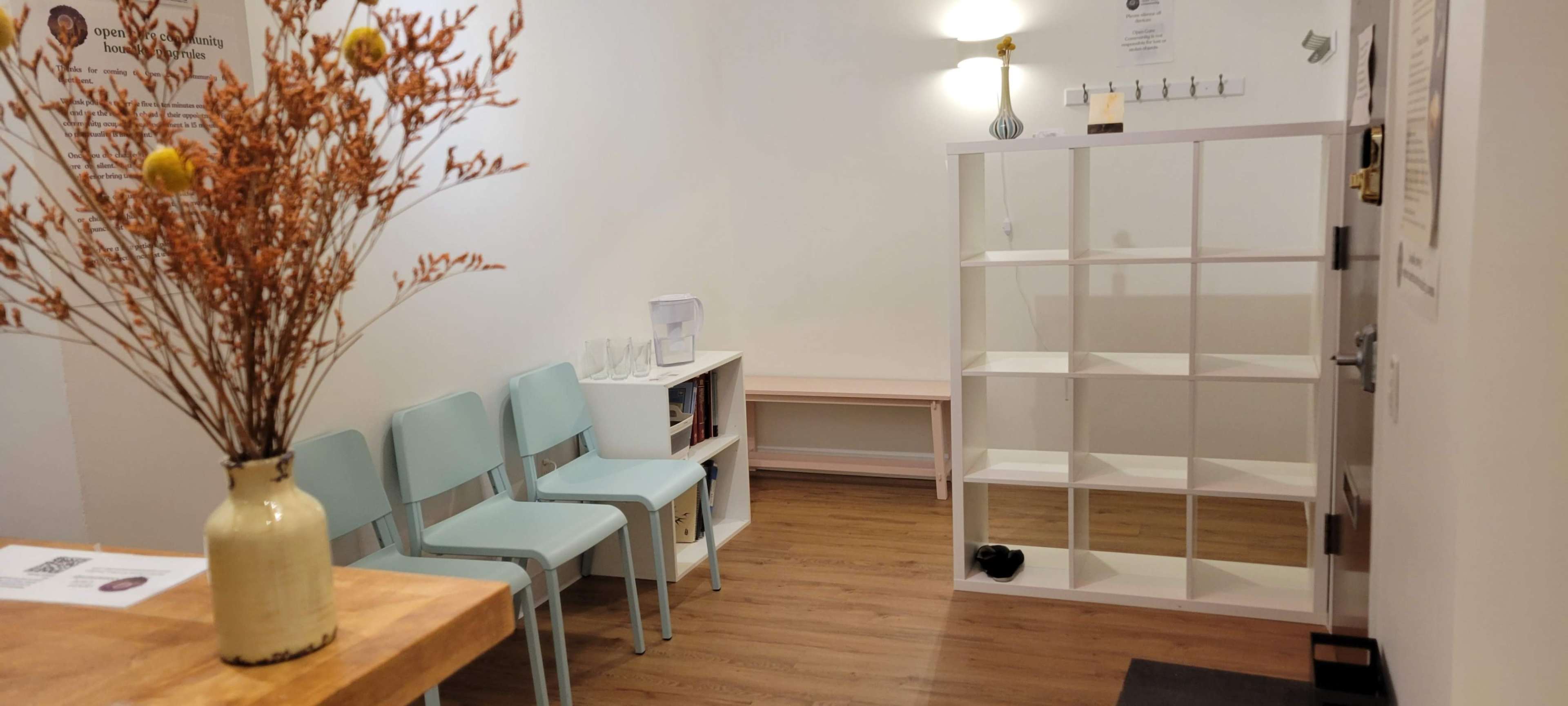 A small waiting room with light blue chairs, a wooden table, a white shelving unit, and a flower arrangement in a vase.