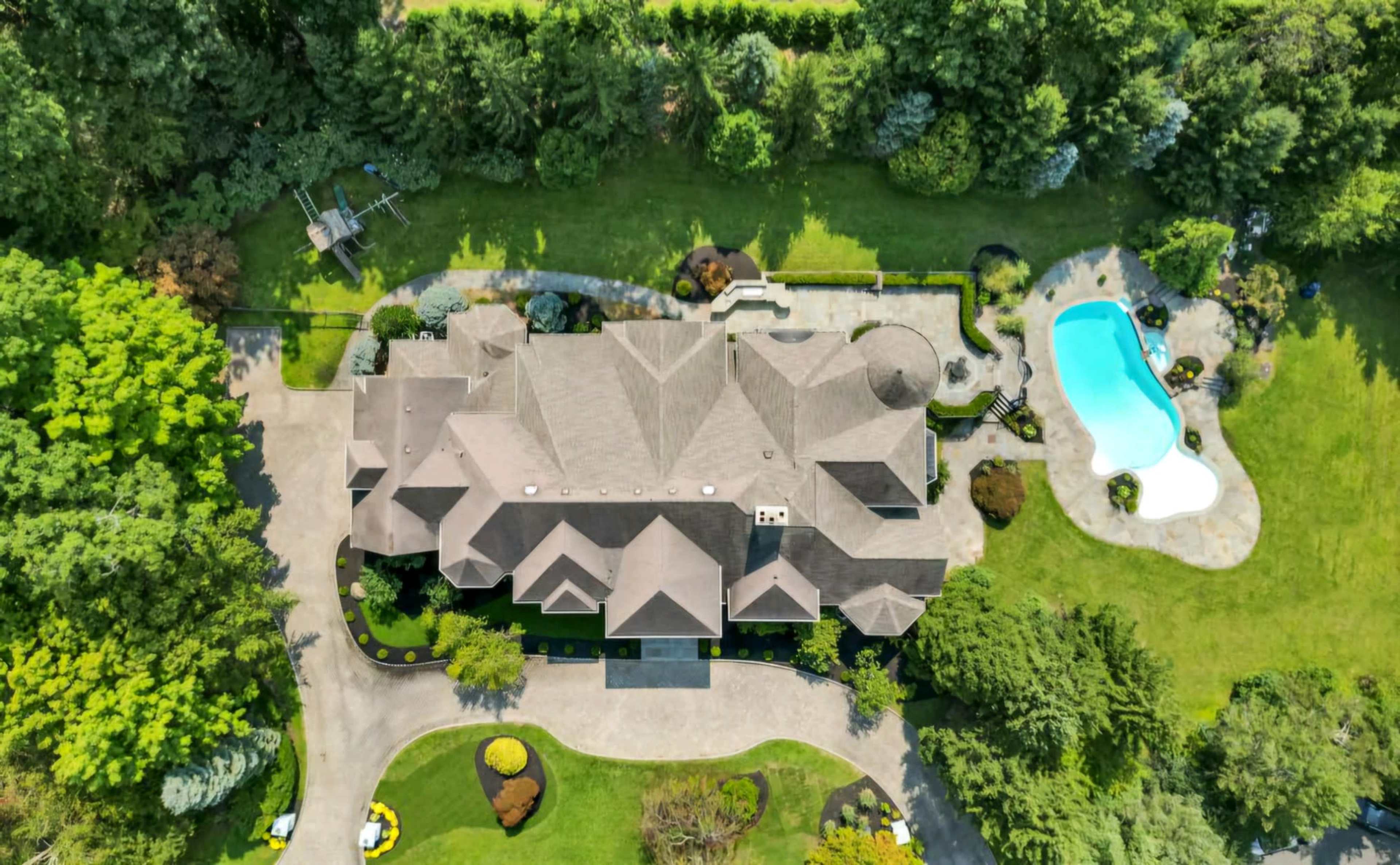 The image shows a large residential home with a complex roof design, surrounded by a well-maintained lawn and a swimming pool.