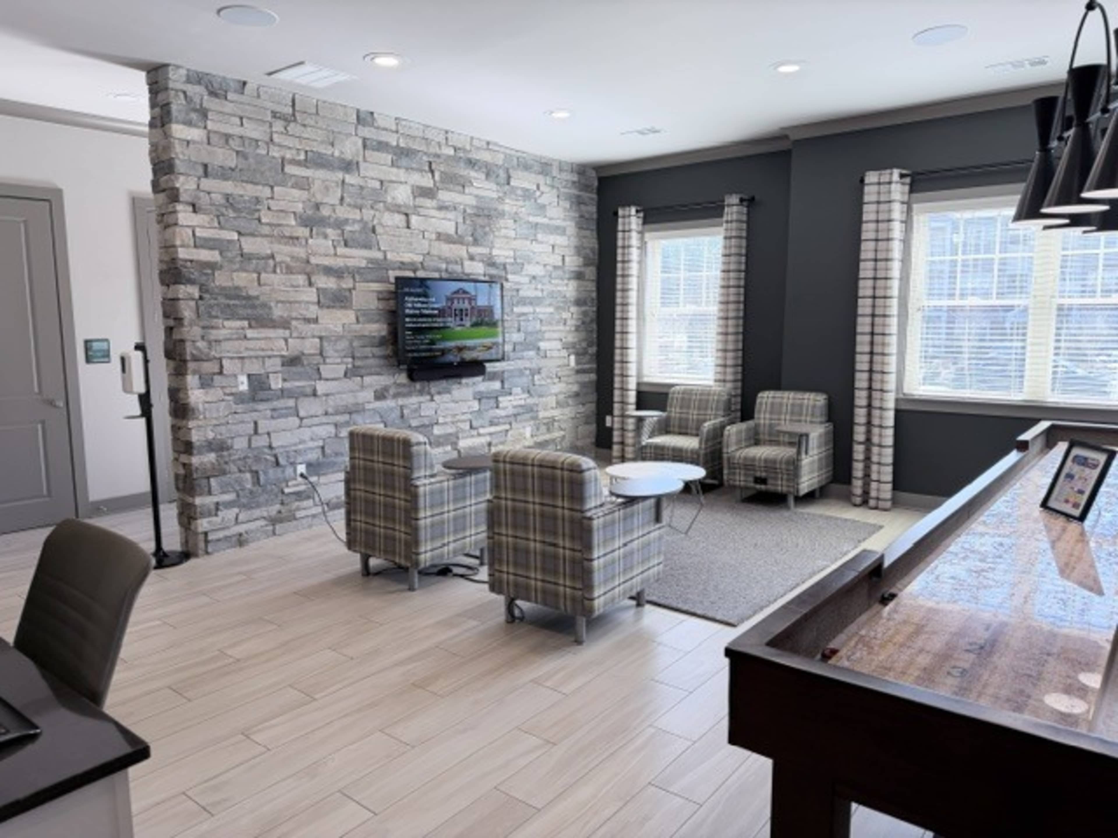 The image shows a modern lounge area with two plaid armchairs, a coffee table, and a television mounted on a stone feature wall, all set against a backdrop of large windows.