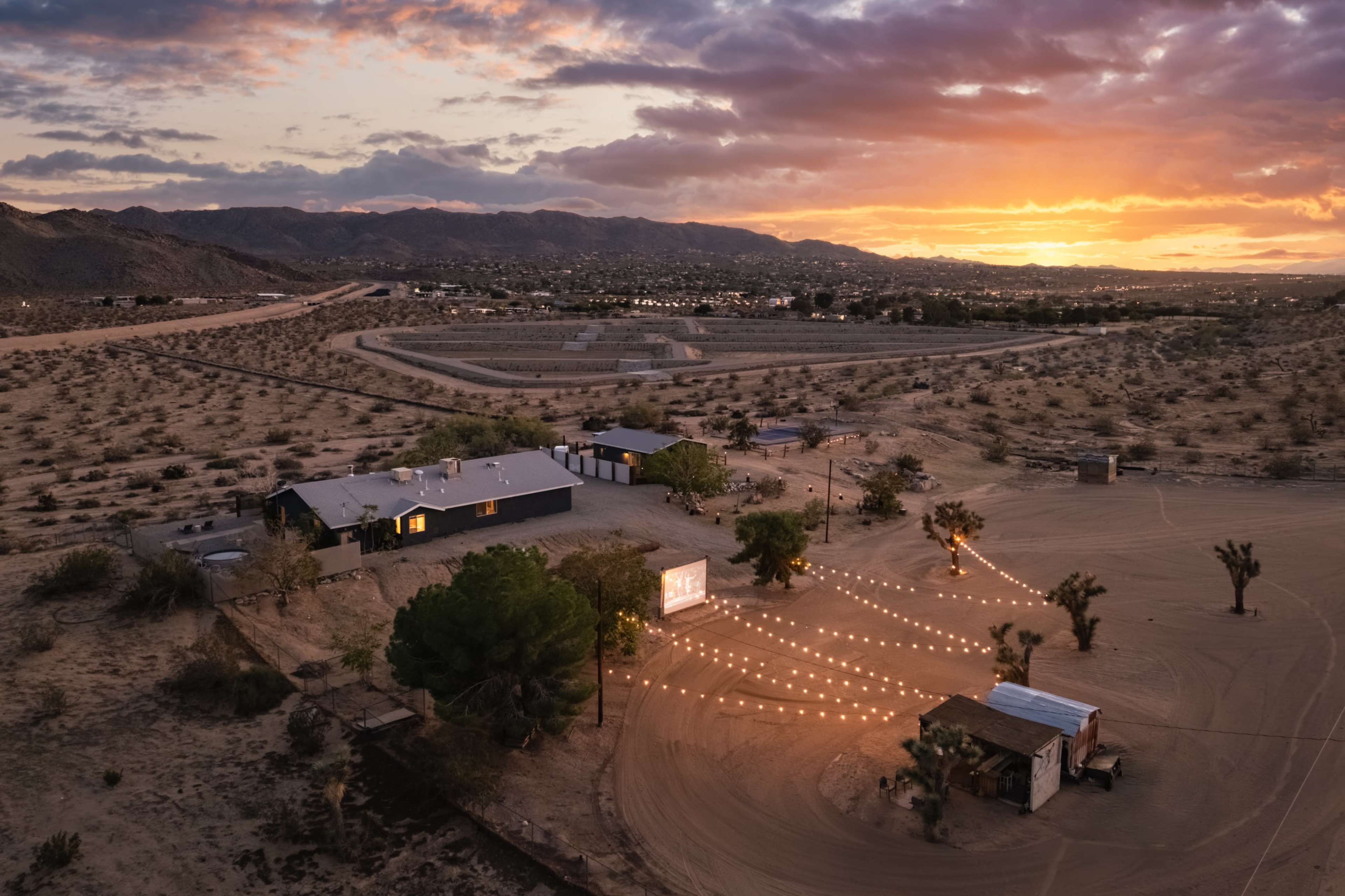 A low-light aerial view shows a desert landscape at sunset, featuring a modern building and string lights illuminating a gathering area surrounded by trees and mountains in the background.