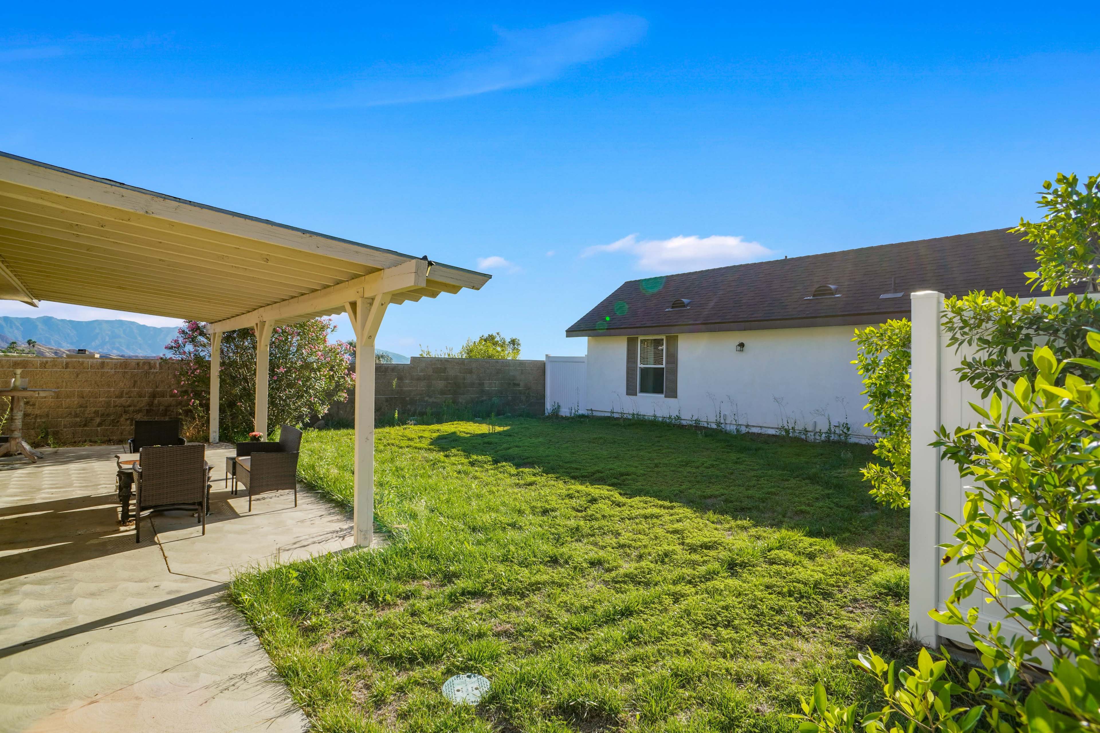 A patio with seating overlooks a grassy backyard surrounded by a fence and bordered by shrubs and a neighboring house.