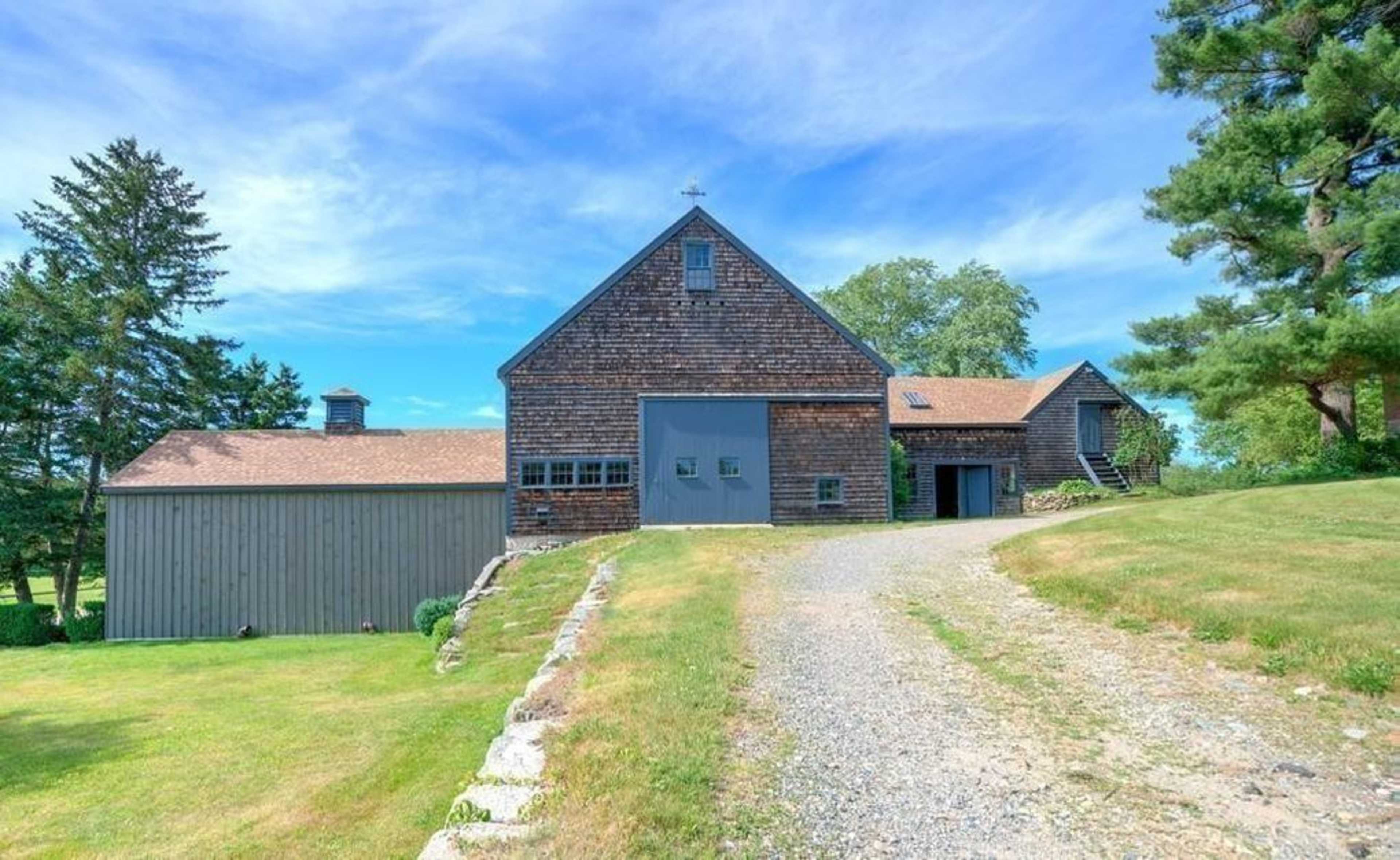A gravel driveway leads to a large wooden barn with a gable roof and a smaller annex beside it, set against a clear blue sky.