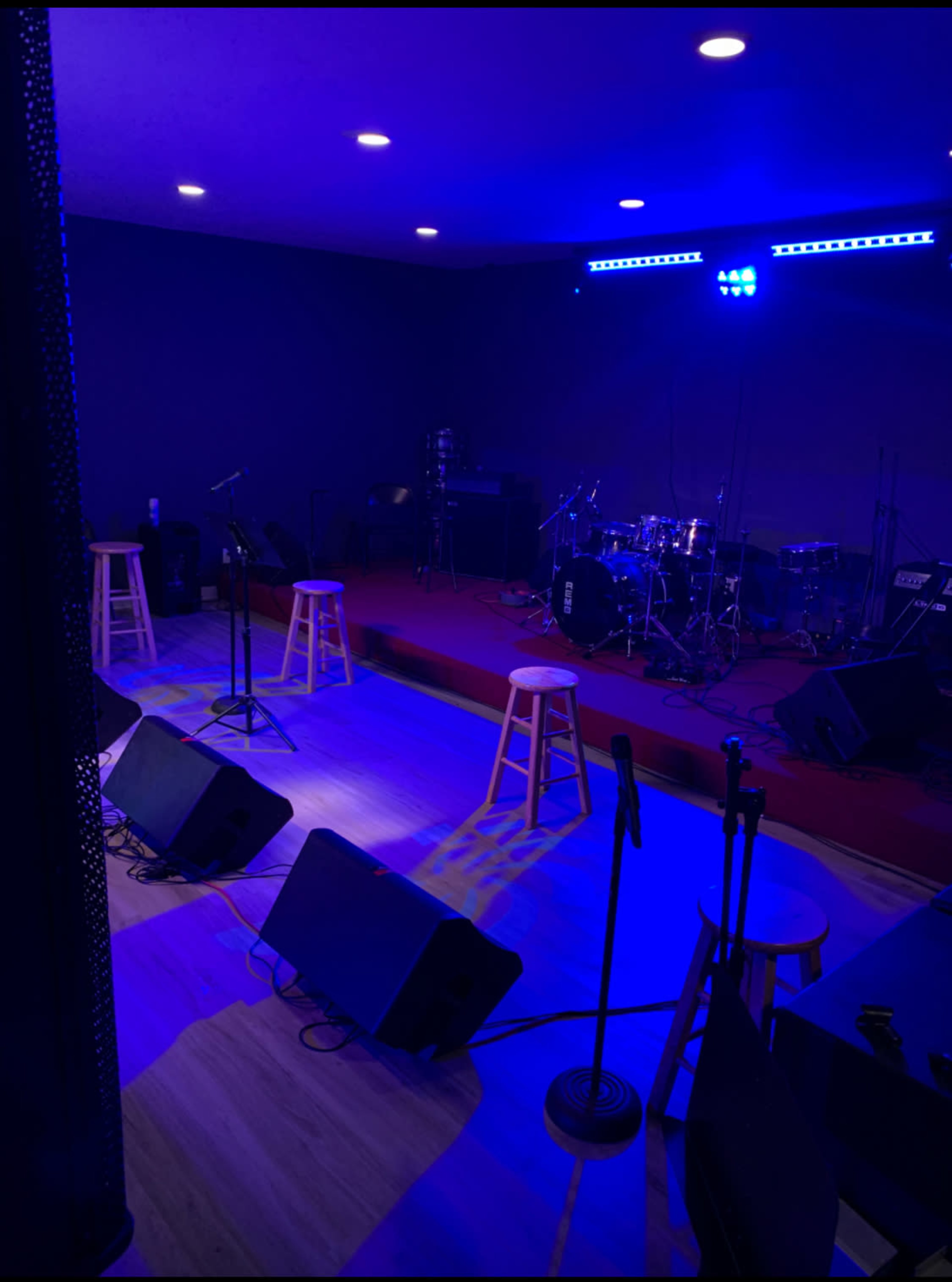 A stage setup with microphones, musical instruments, and stool seating is illuminated by blue lights.
