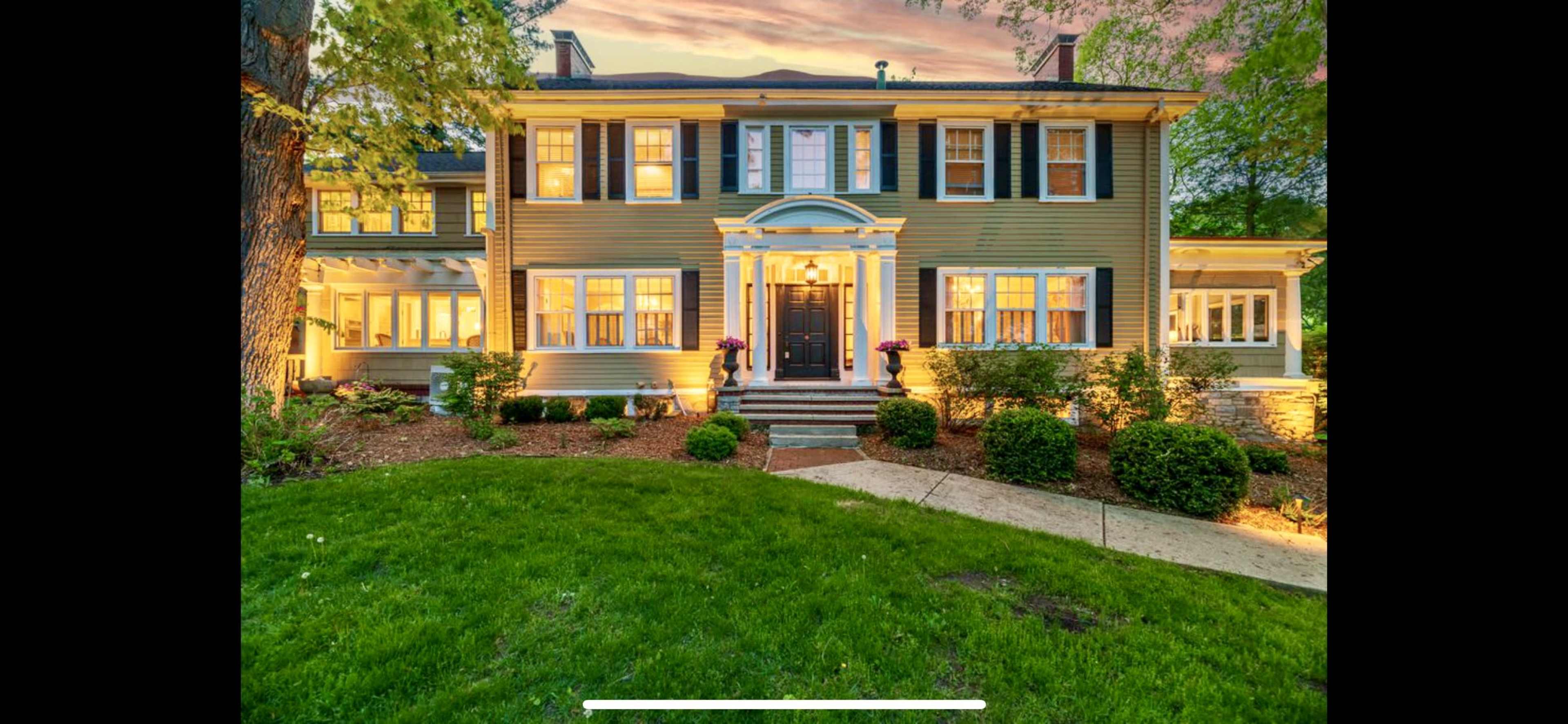 A two-story house with light green siding, white trim, and well-landscaped gardens illuminated by evening light.