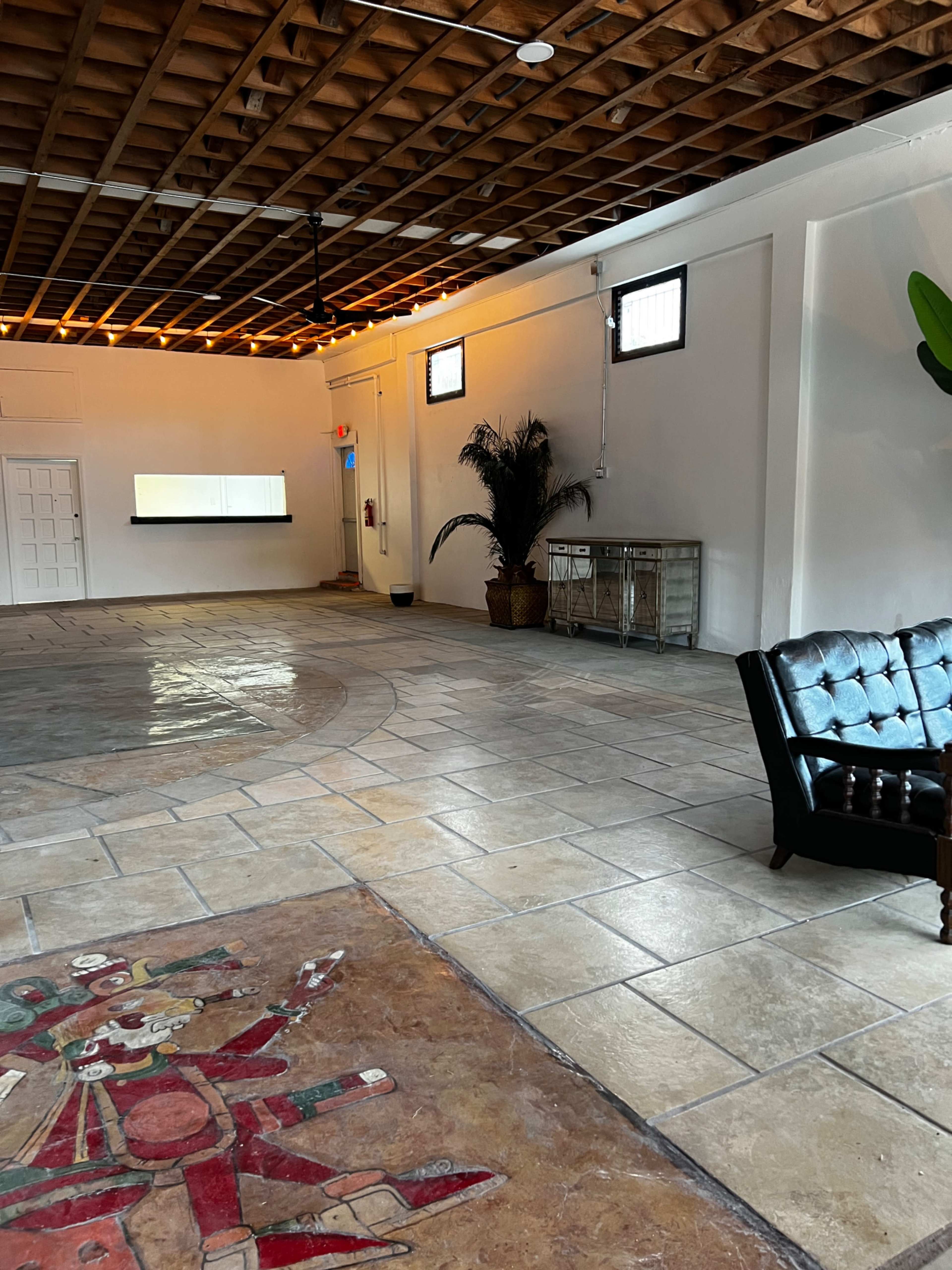 A spacious room with a tiled floor, featuring a dark leather sofa, a decorative mural on the floor, and a few potted plants along the walls.