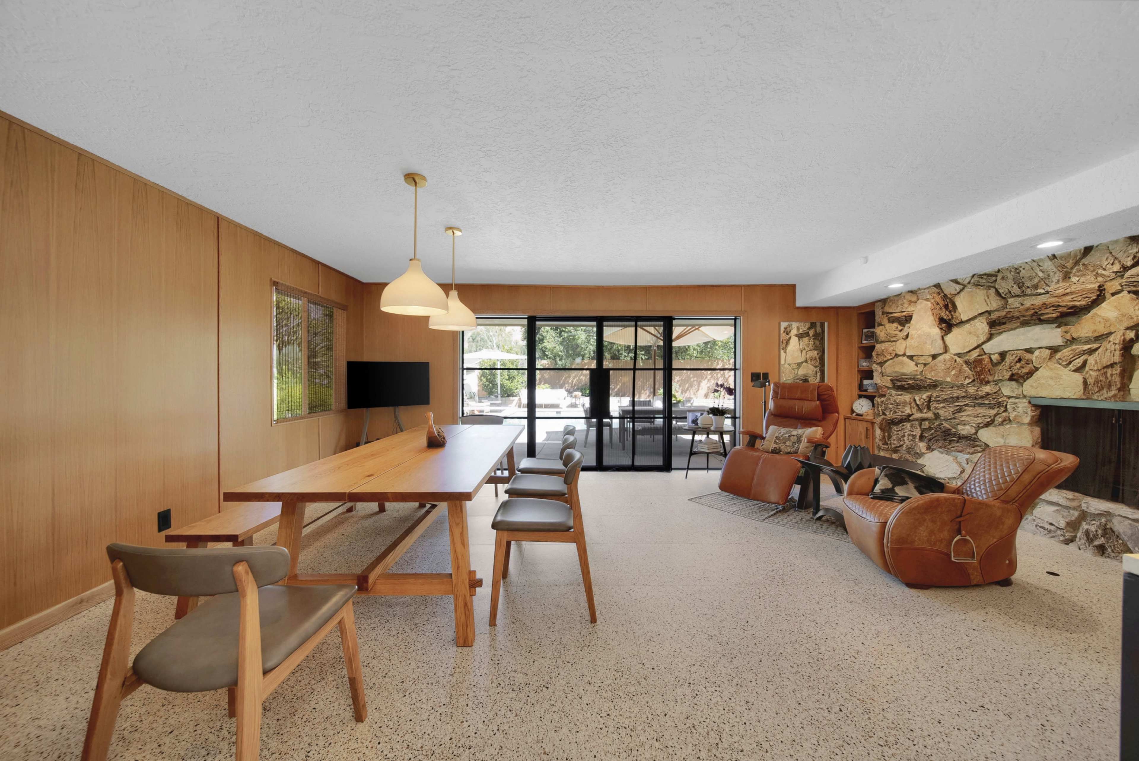 The image shows a modern living area featuring a wooden dining table, lounge chairs, a stone accent wall, and large windows overlooking an outdoor space.