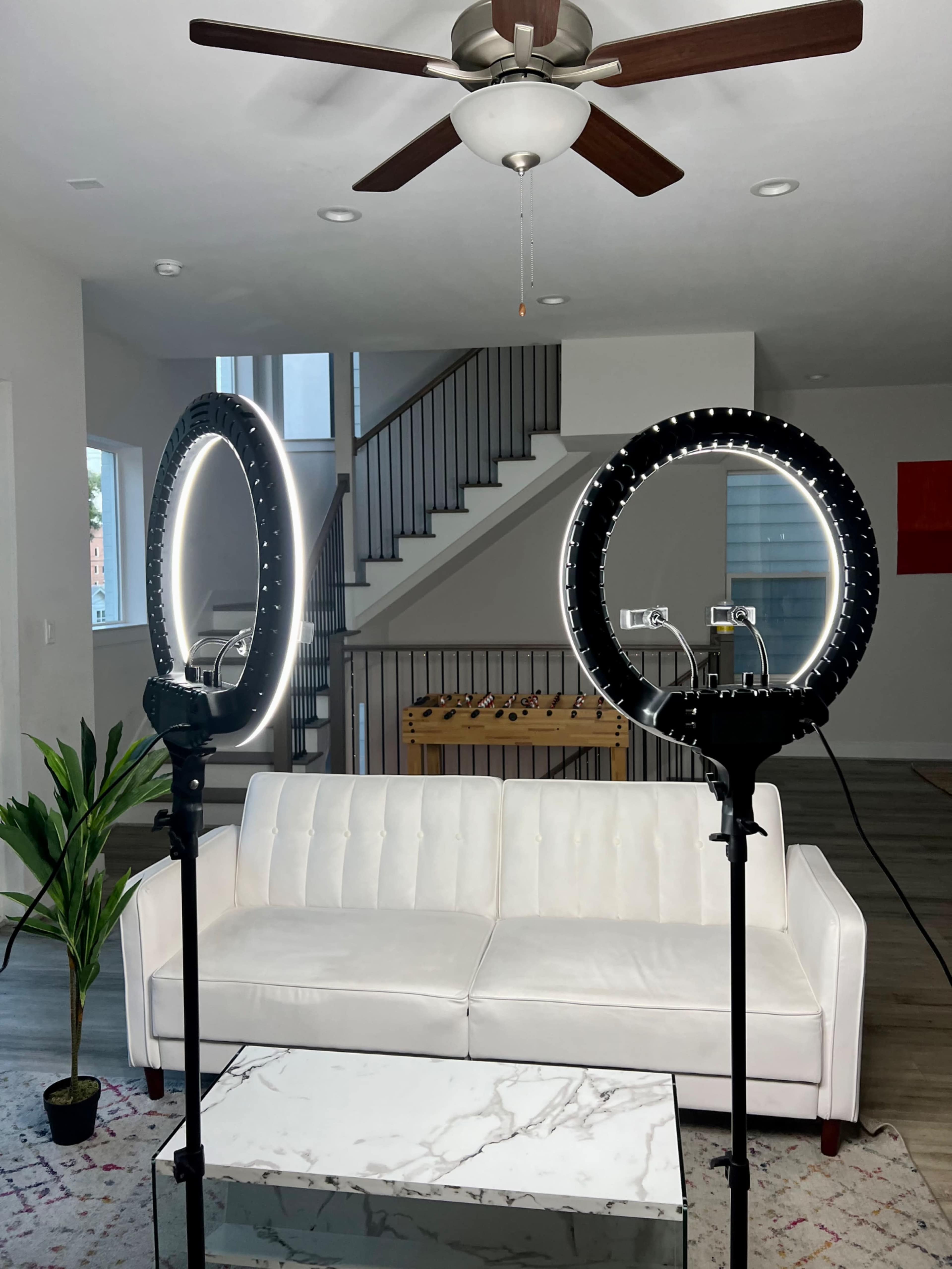Two ring lights positioned on stands in a living room with a white sofa, a glass coffee table, and a staircase in the background.