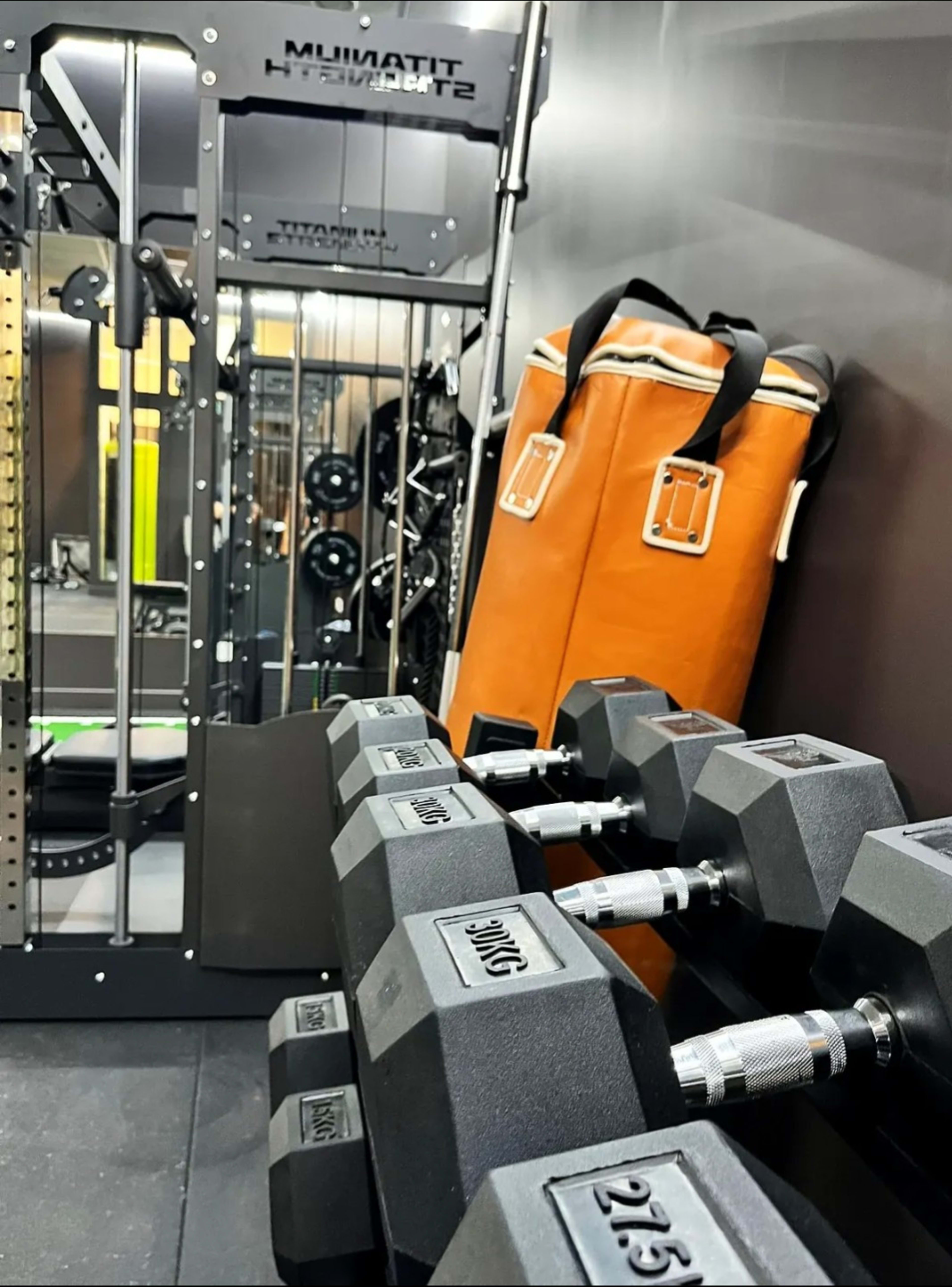 The image shows a home gym setup featuring a set of dumbbells, strength training equipment, and an orange duffel bag.