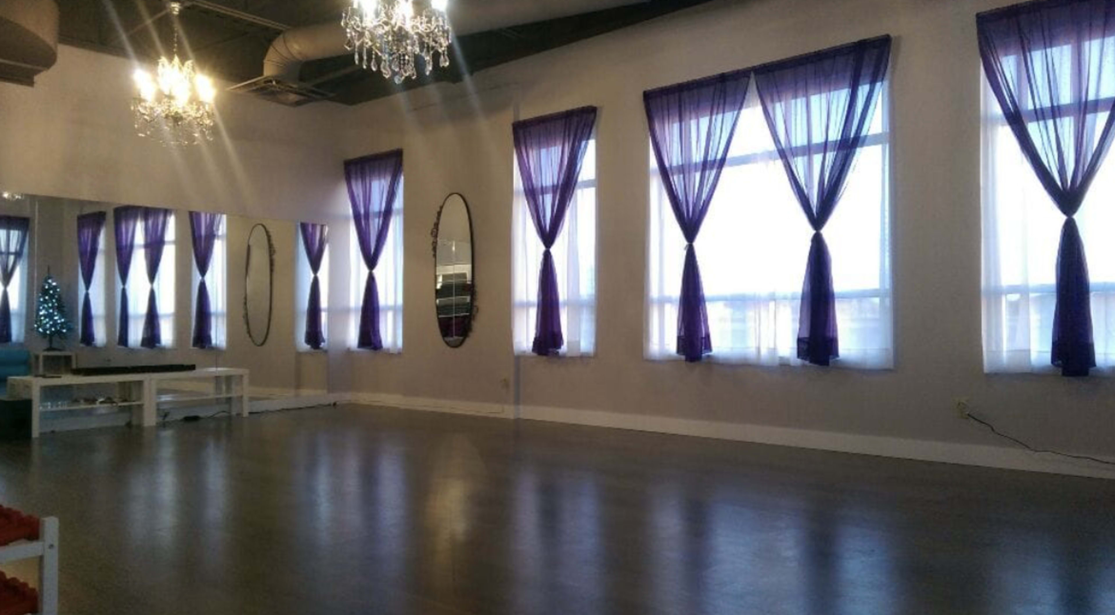 A spacious studio with large windows draped in purple curtains and several mirrors reflecting the natural light.