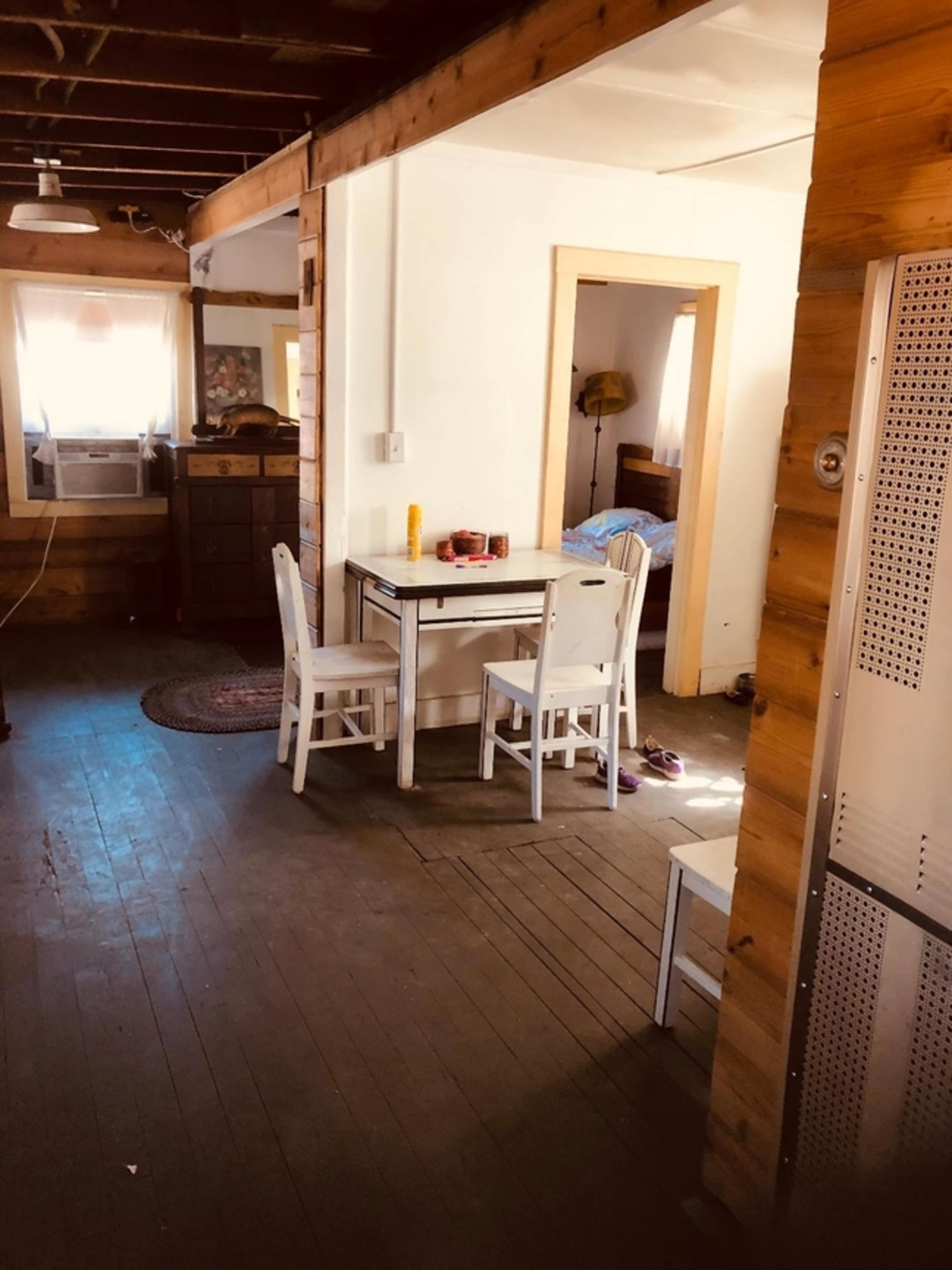 A simple interior space with a small table and two chairs, a wooden floor, and a doorway leading to another room.
