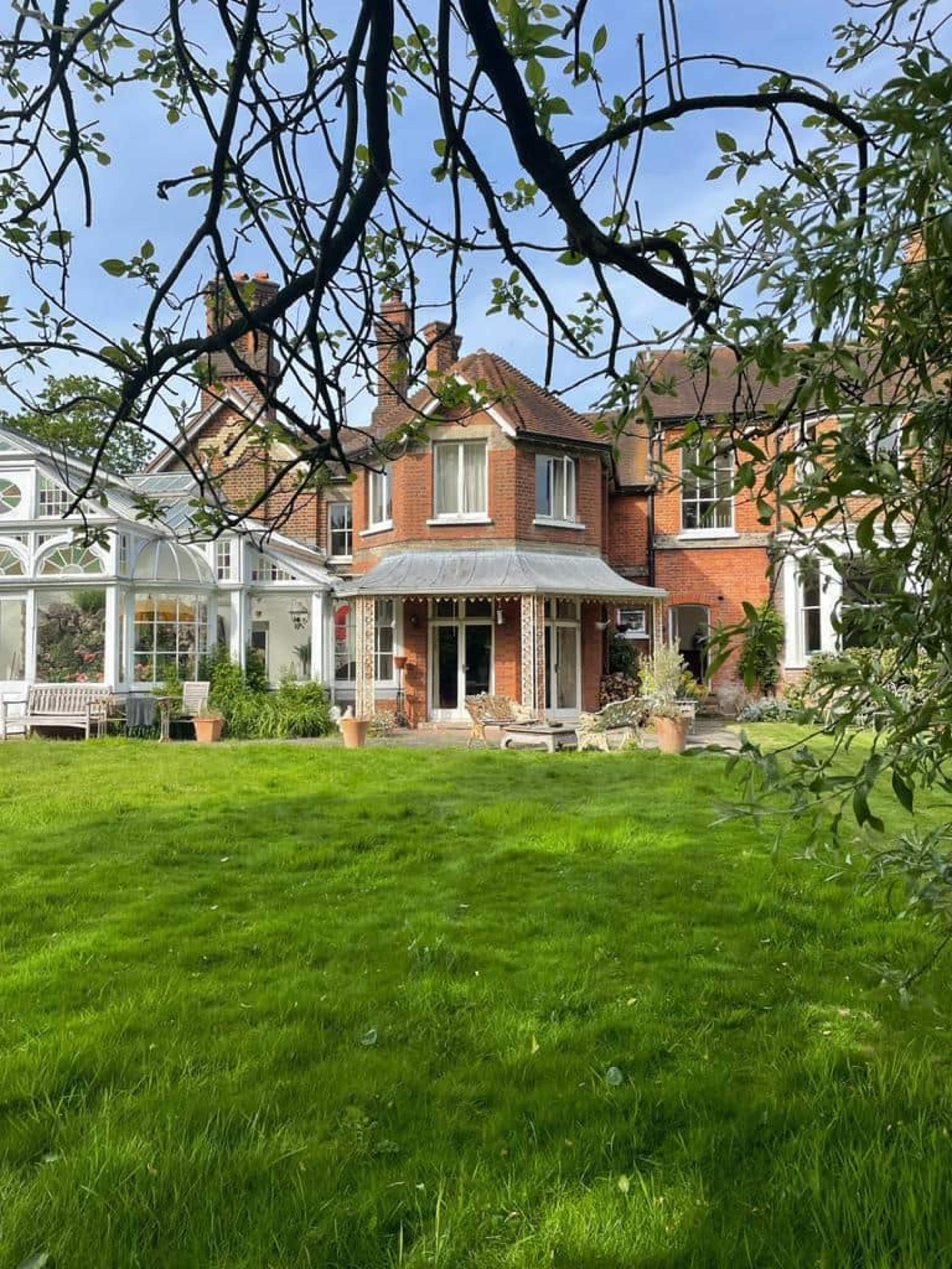 A brick house with multiple sections, including a circular turret, is set in a green lawn surrounded by trees and plants.