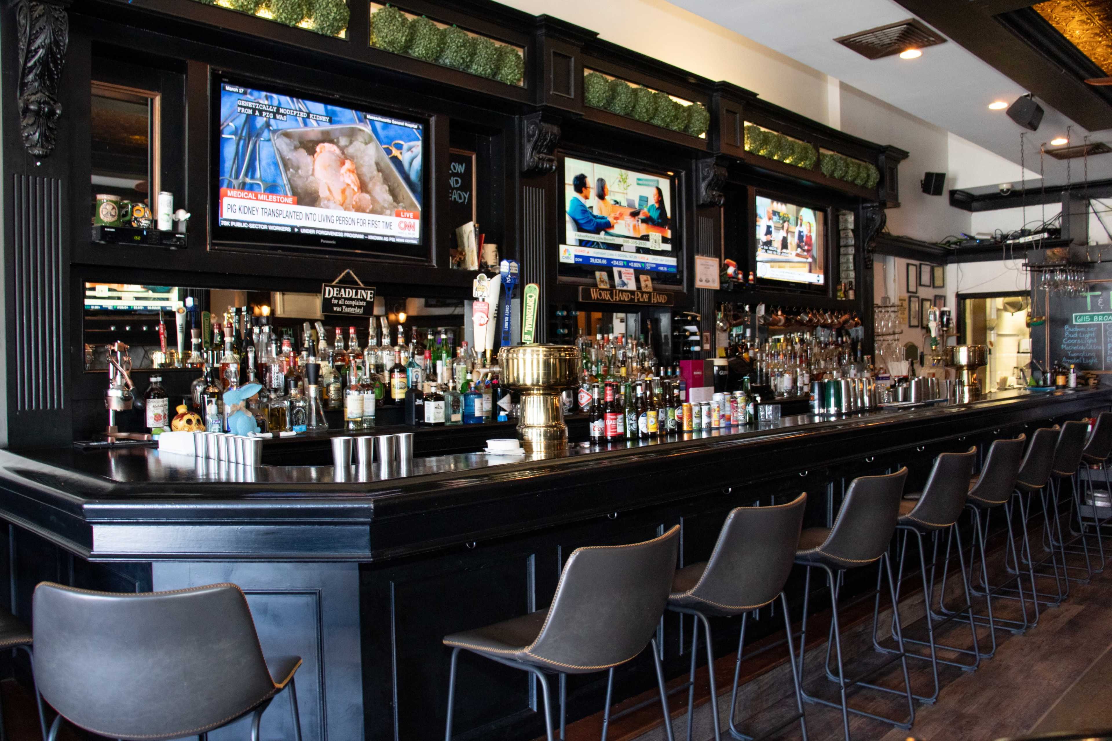 A bar with a long counter, multiple television screens displaying news, and a variety of bottles lined up on the shelves behind the bar.