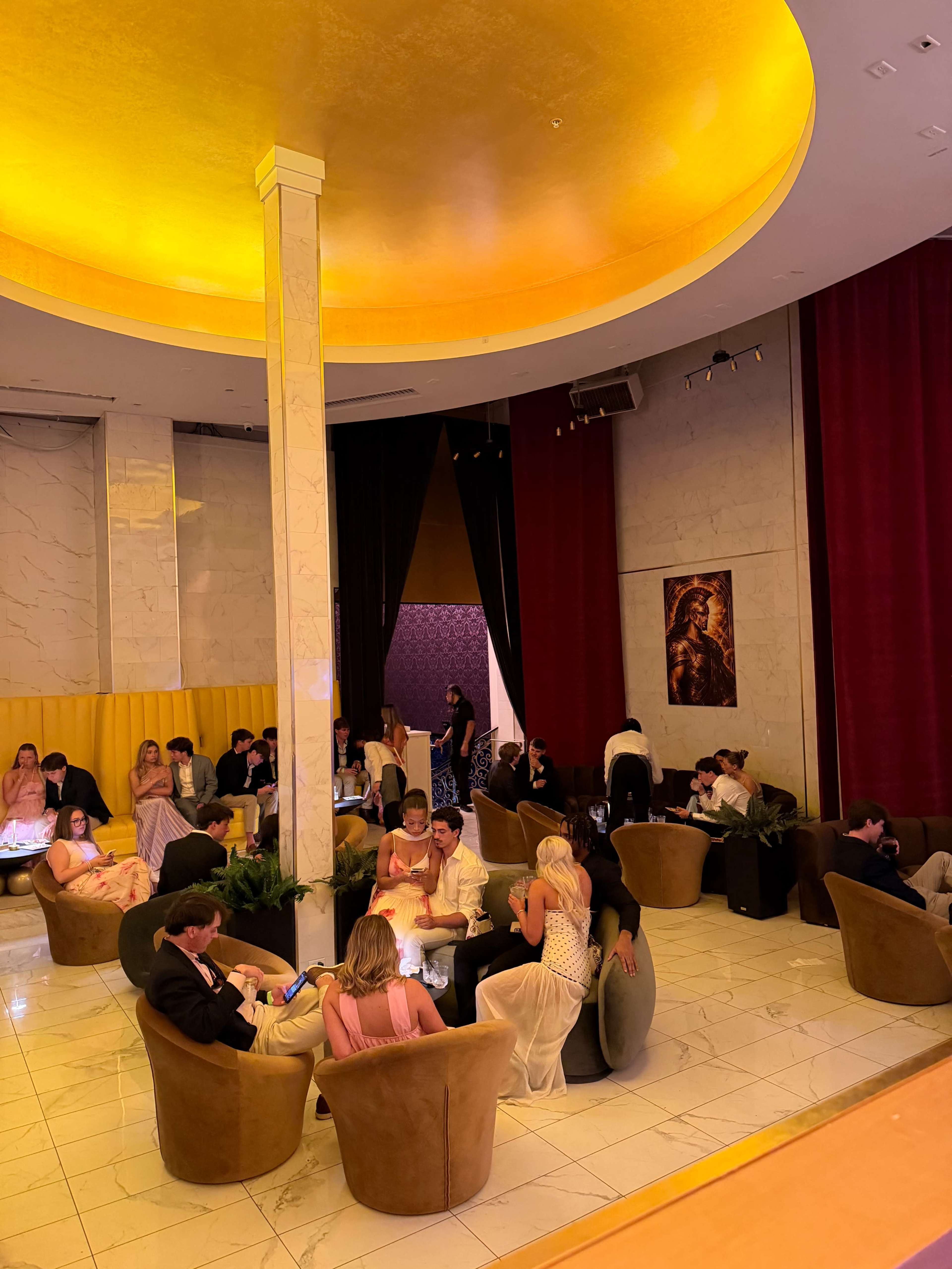 A group of elegantly dressed people socializes in a stylish lounge featuring plush seating and ambient lighting.