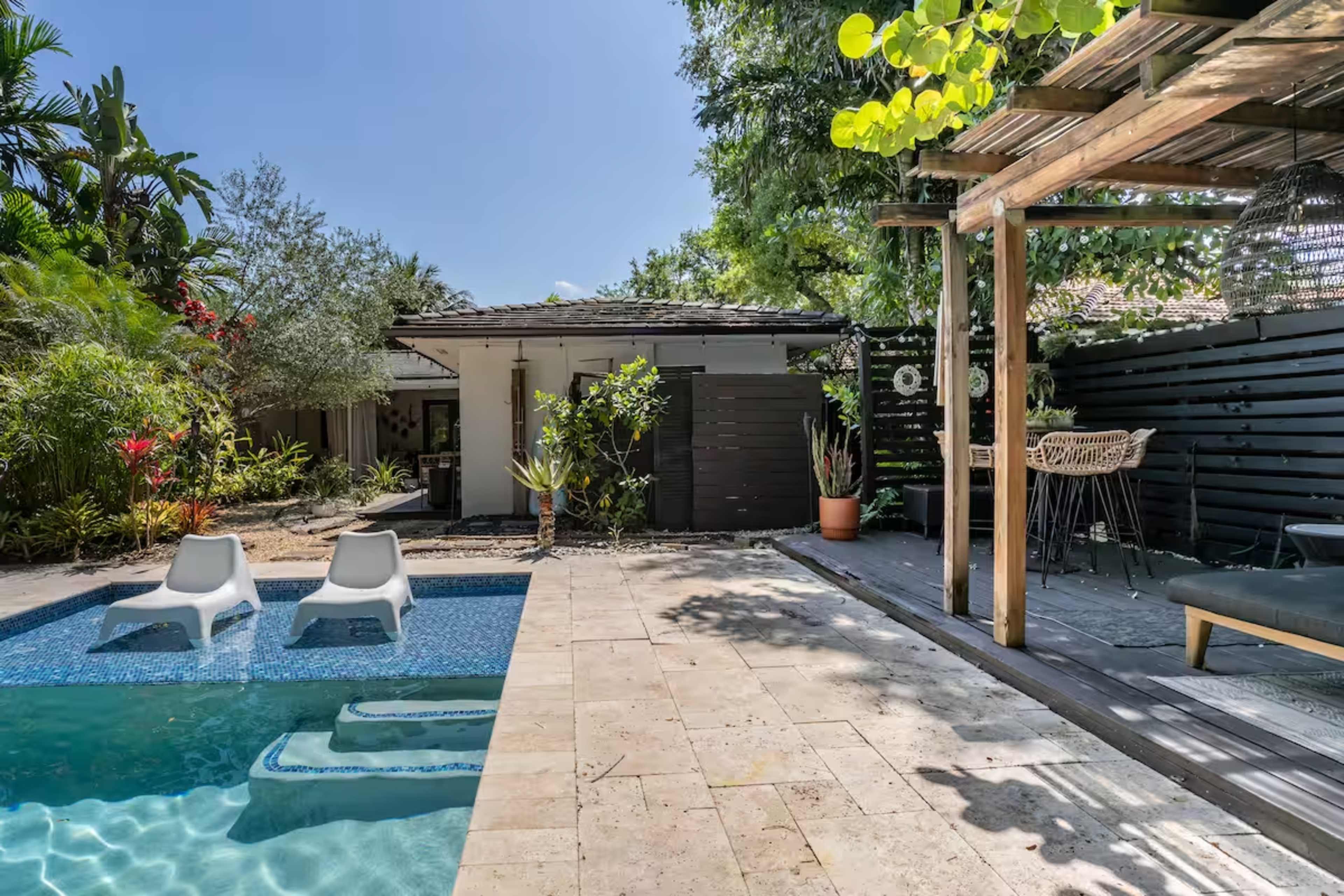 The image shows a backyard with a swimming pool, two white lounge chairs, and a seating area under a wooden pergola surrounded by various plants and landscaping.