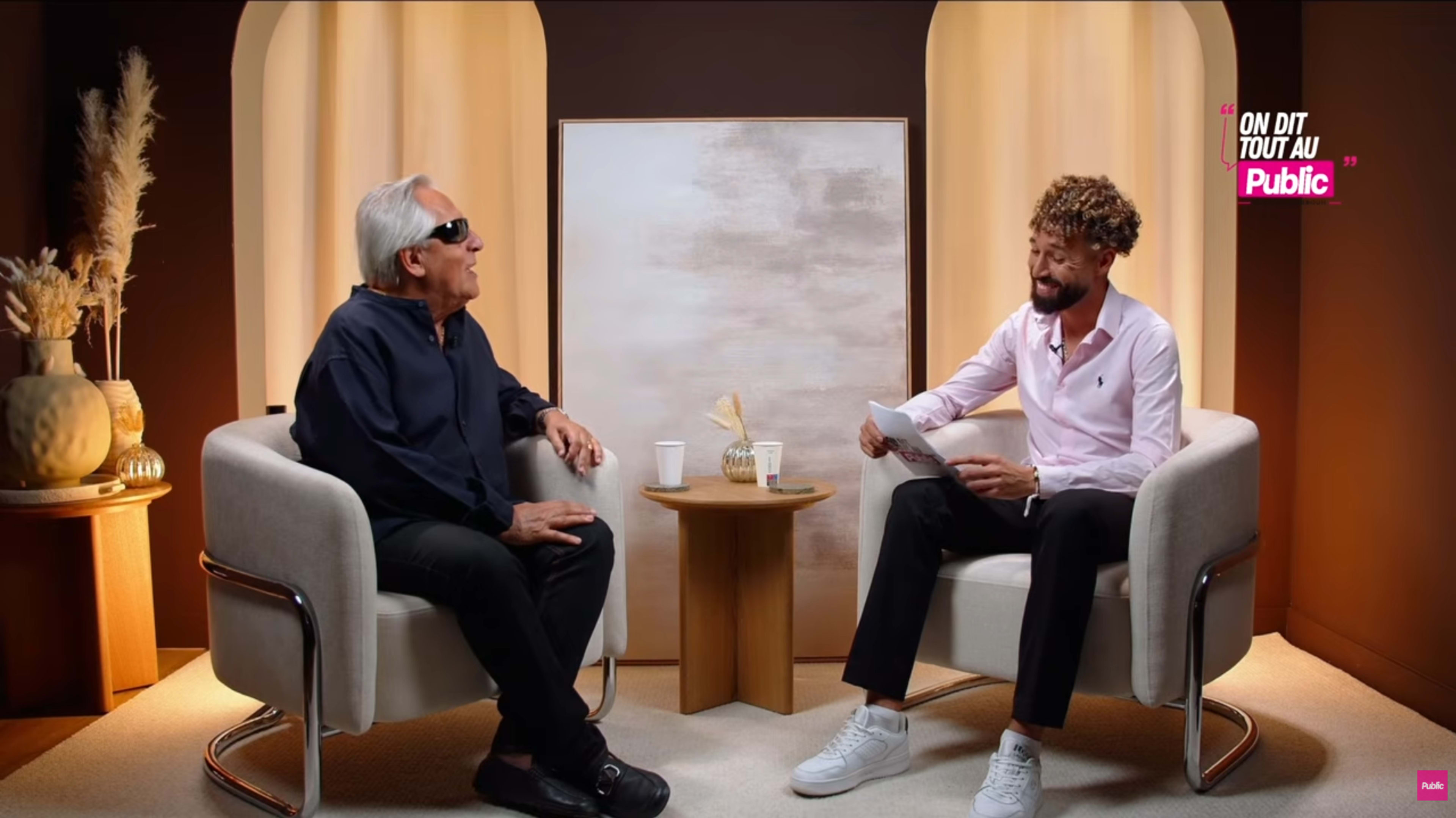 Two men are seated in modern armchairs, engaged in conversation, with a decorative backdrop featuring a large abstract painting and a small table holding a cup and decorative items.