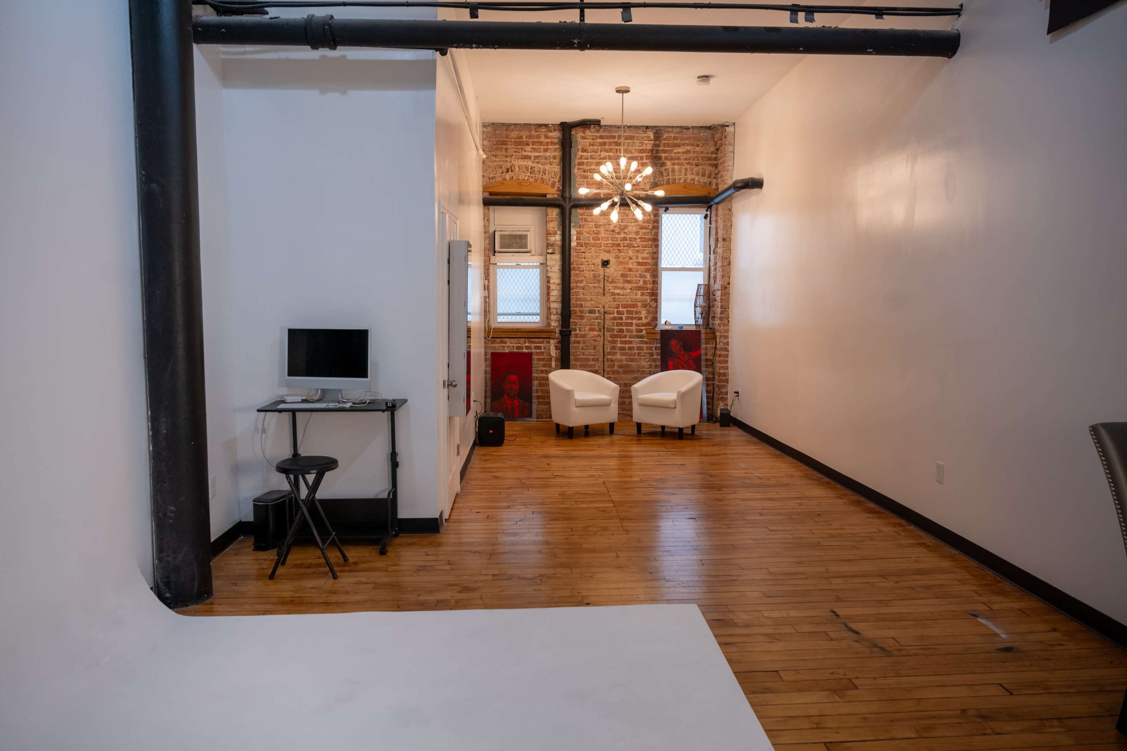 The image shows a spacious, modern room featuring hardwood flooring, exposed brick walls, two white chairs at the far end, and a desk with a computer near the entrance.