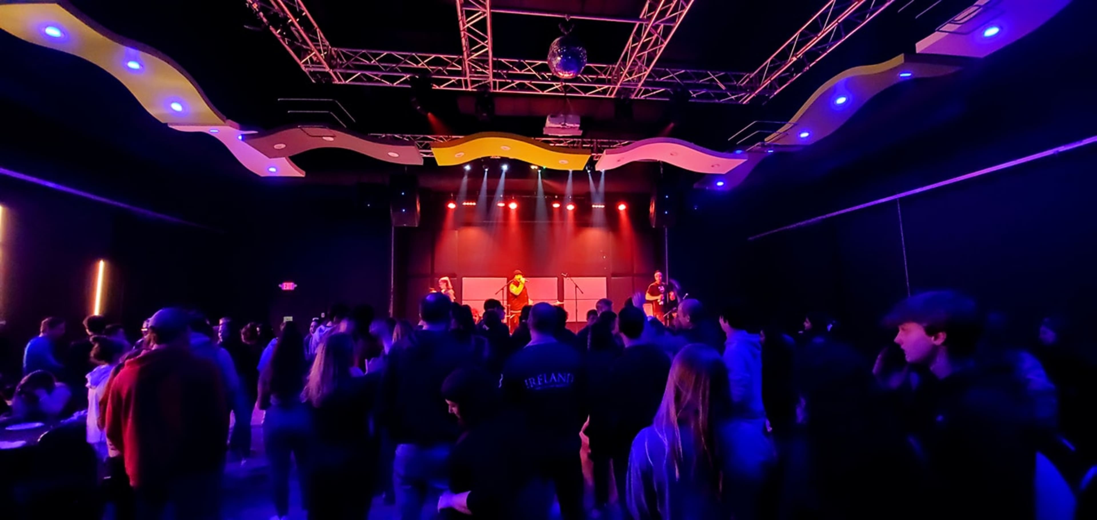 A band performs on stage in a dimly lit venue filled with an audience.