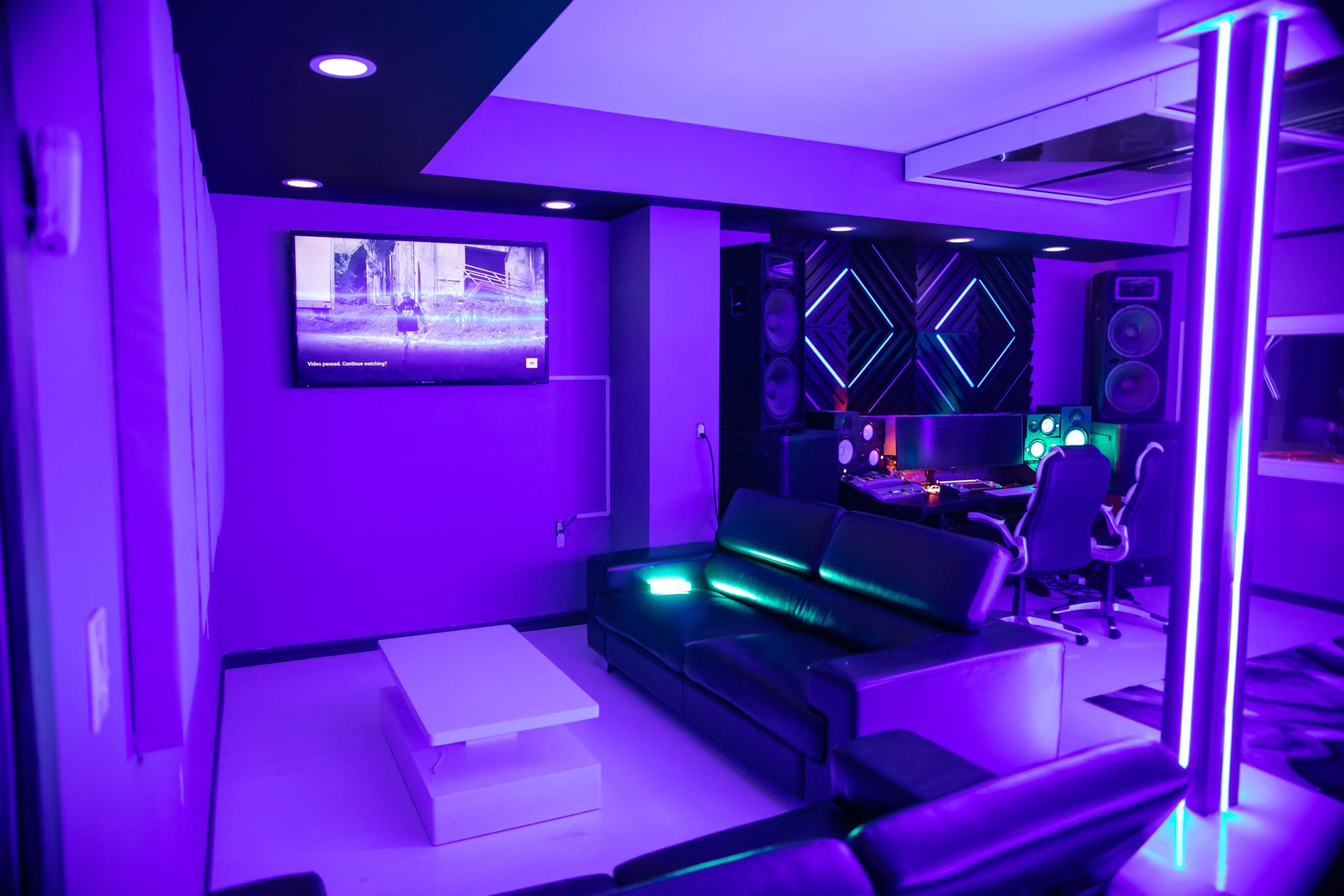 A modern media room with purple lighting, a black sofa, a coffee table, and a workspace featuring audio equipment and a television on the wall.